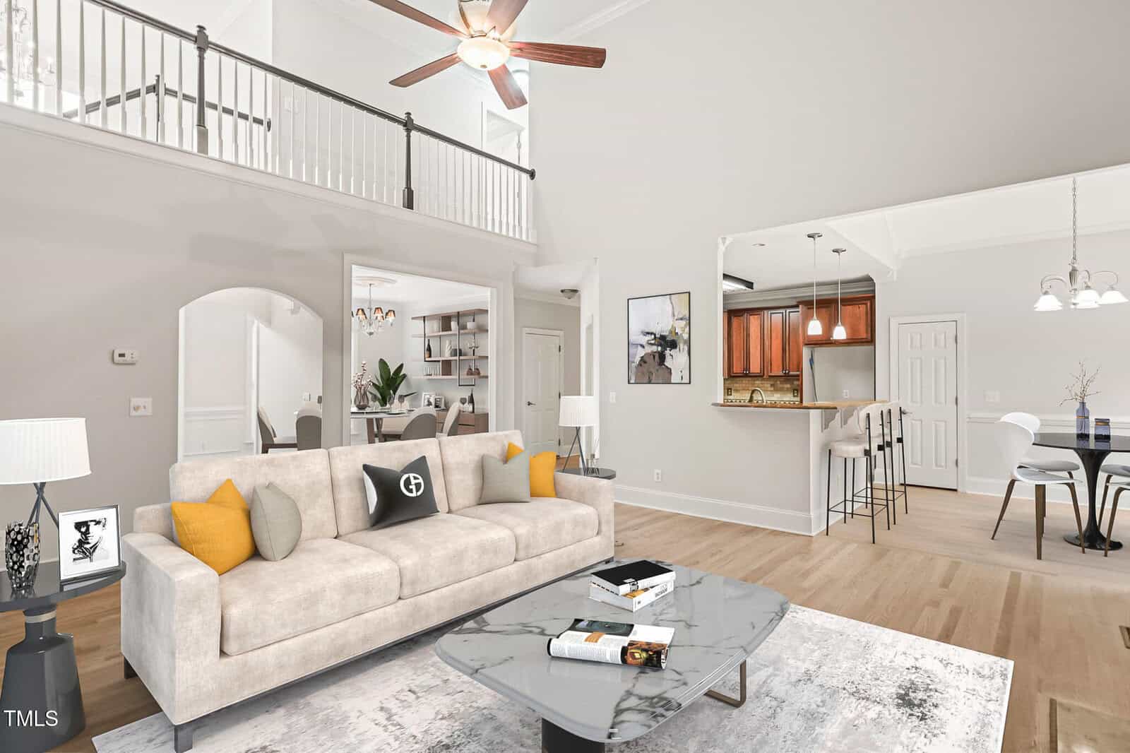 A modern open living area features a beige sofa with accent pillows, a marble coffee table, a kitchen with a breakfast bar, and a dining area. A balcony overlooks the space, and neutral walls and wood floors complete the room.