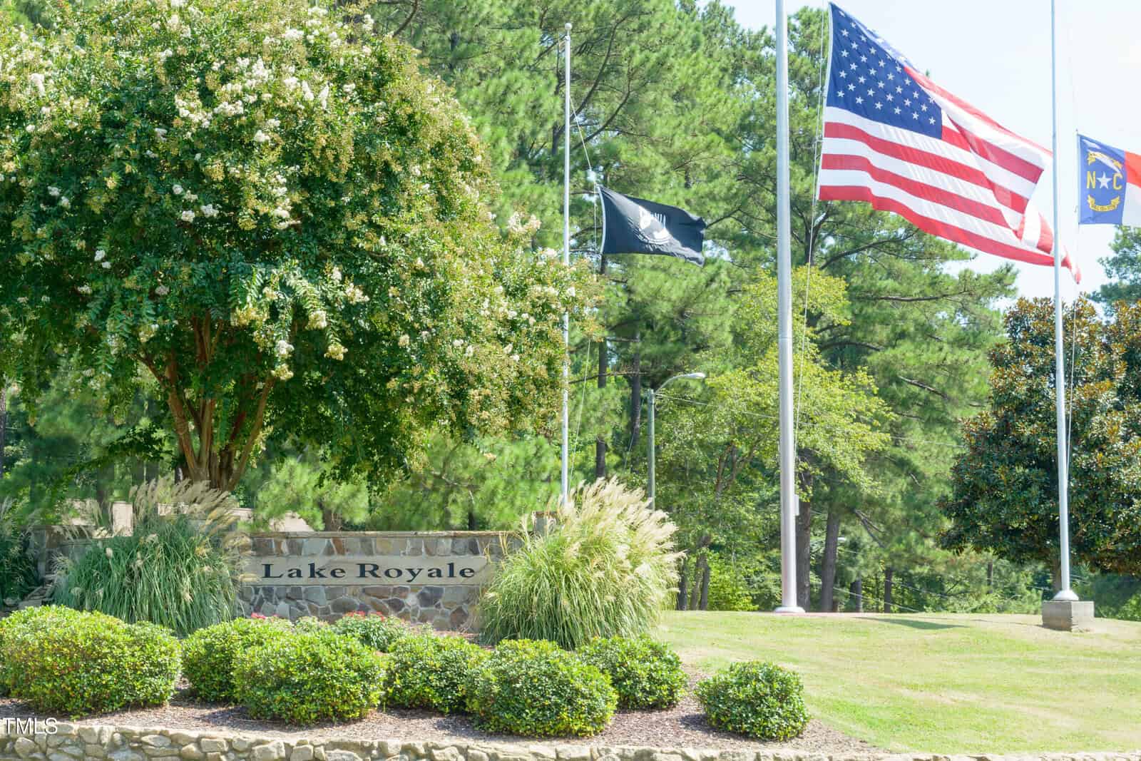 A stone sign reading “Lake Royale” sits by trimmed bushes and a tree. Behind it, three flagpoles display the American flag, North Carolina flag, and a black flag, with pine trees in the background.