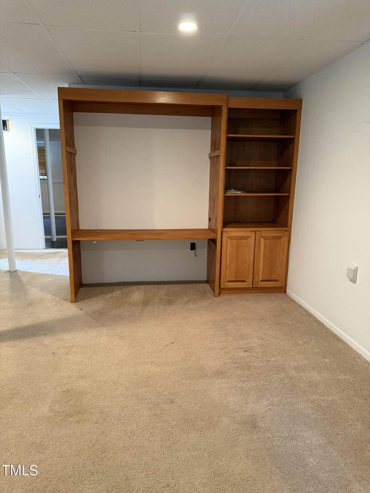 Built-in wooden shelving unit with open desk space, four shelves, and two closed cabinets, set against a white wall in a carpeted room with white ceiling tiles and recessed lighting.