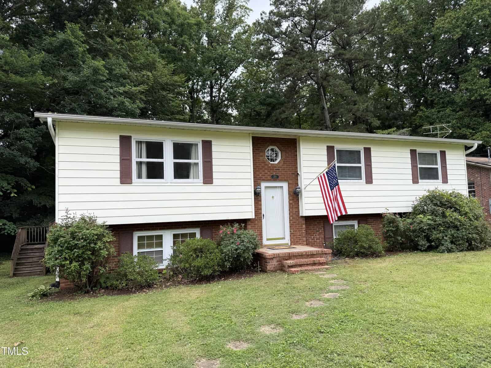 A split-level house with cream siding and red shutters sits on a grassy lawn at 210 Crest Road in Henderson. There are bushes in front, brick steps leading to the main door, and an American flag mounted next to the entrance. Trees are in the background.