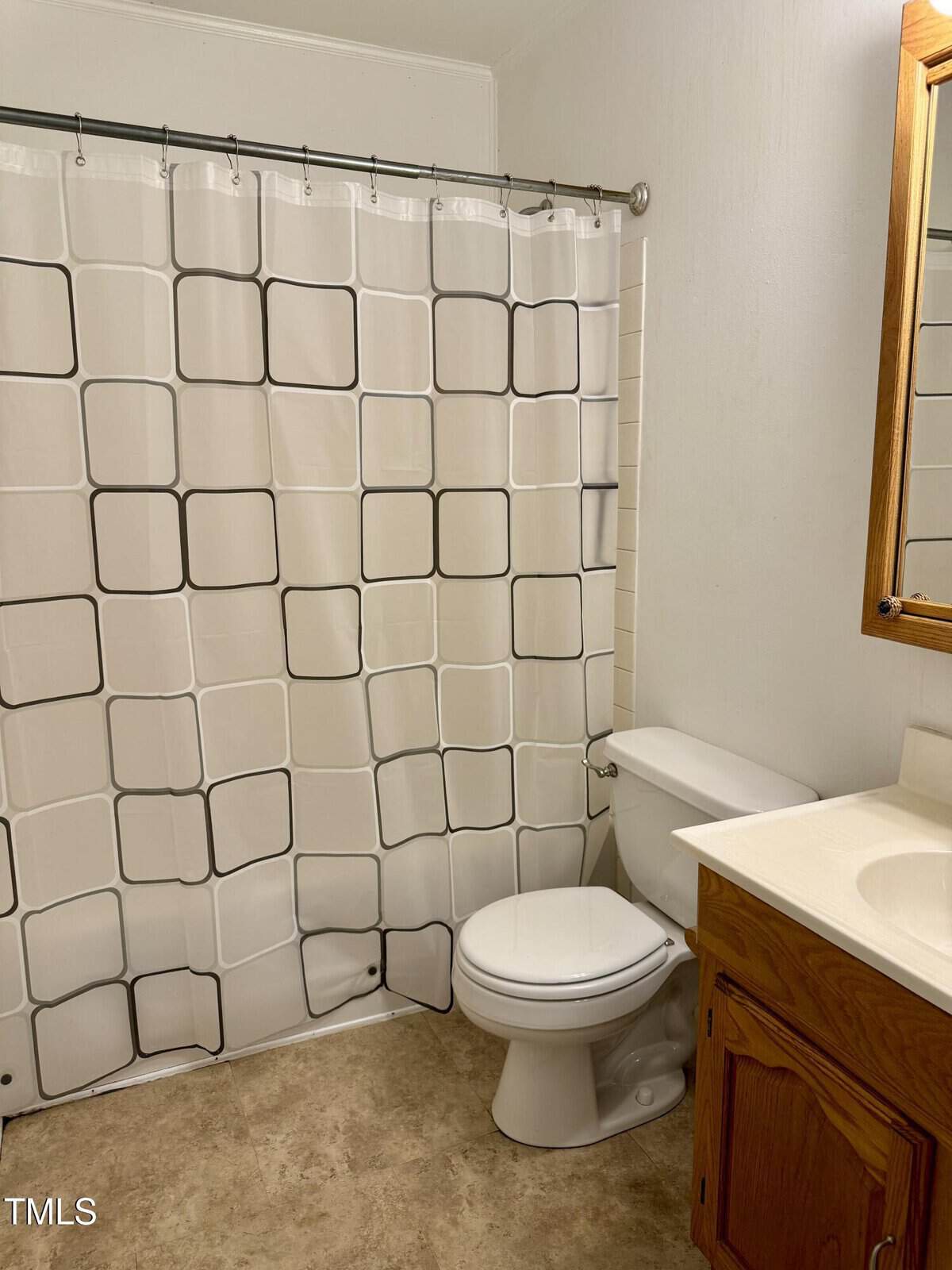 A bathroom with a white shower curtain featuring a black square pattern, a white toilet, a wooden vanity with a sink, a mirror above the sink, and light-colored flooring.