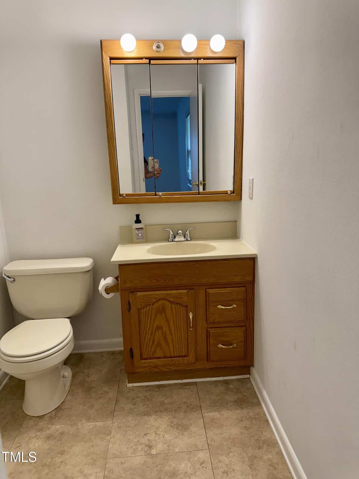 A small bathroom with a toilet on the left, a wooden vanity with a sink and mirror above it, and three lights above the mirror. The walls are white and the floor has beige tiles.