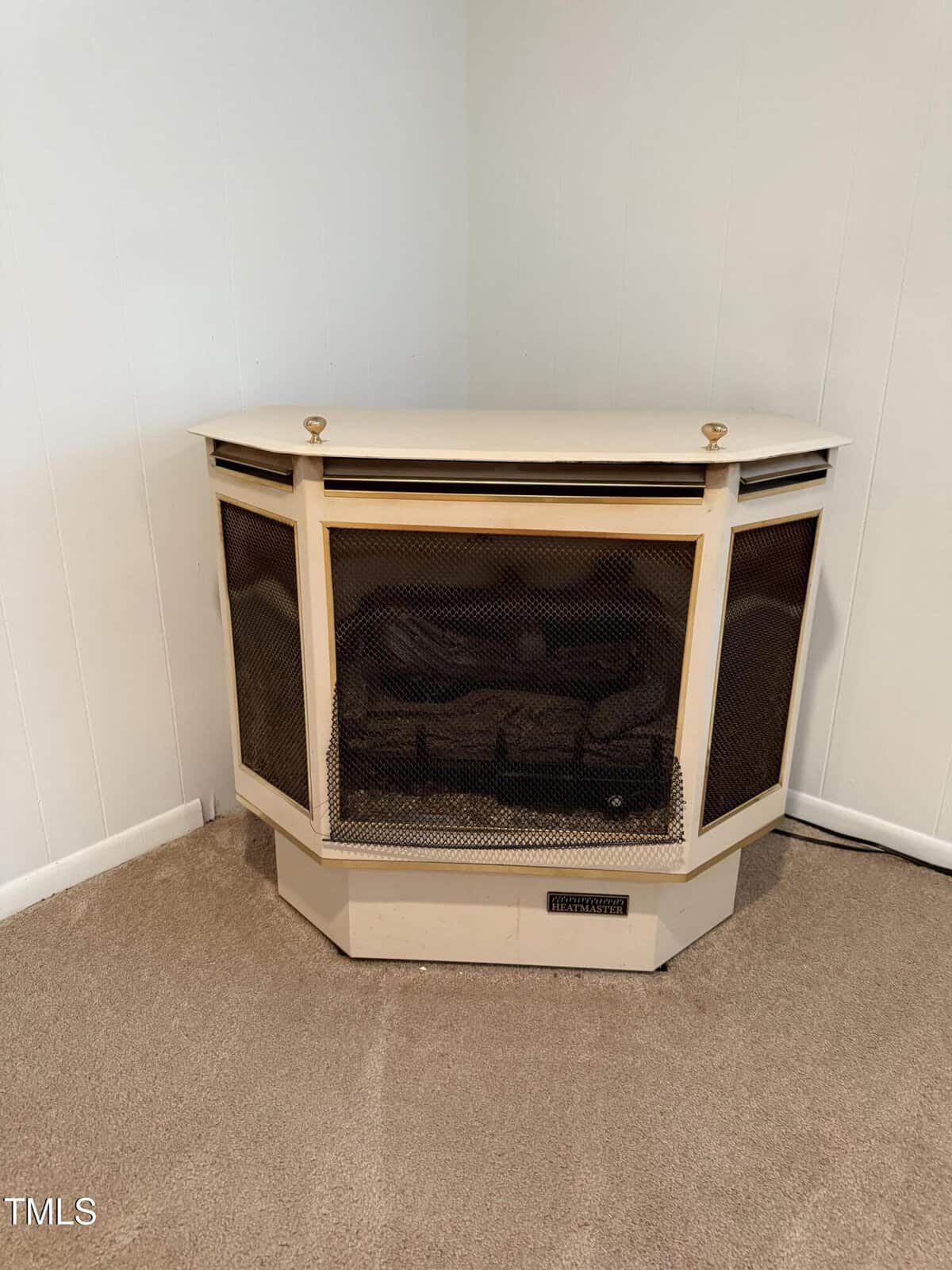 A cream-colored gas fireplace with black mesh screen is set in the corner of a carpeted room with white walls. Two gold knobs are on top, and the unit has a vent at the bottom.
