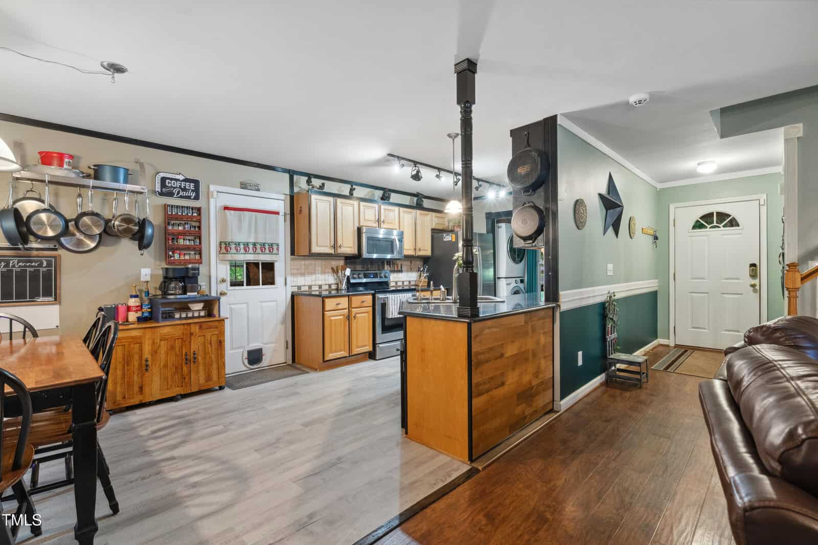Cozy kitchen and entryway with wood cabinets, stainless steel appliances, open shelving, and hanging pots. Green and white walls, wooden floors, dining table, and a white front door with a sidelight window.