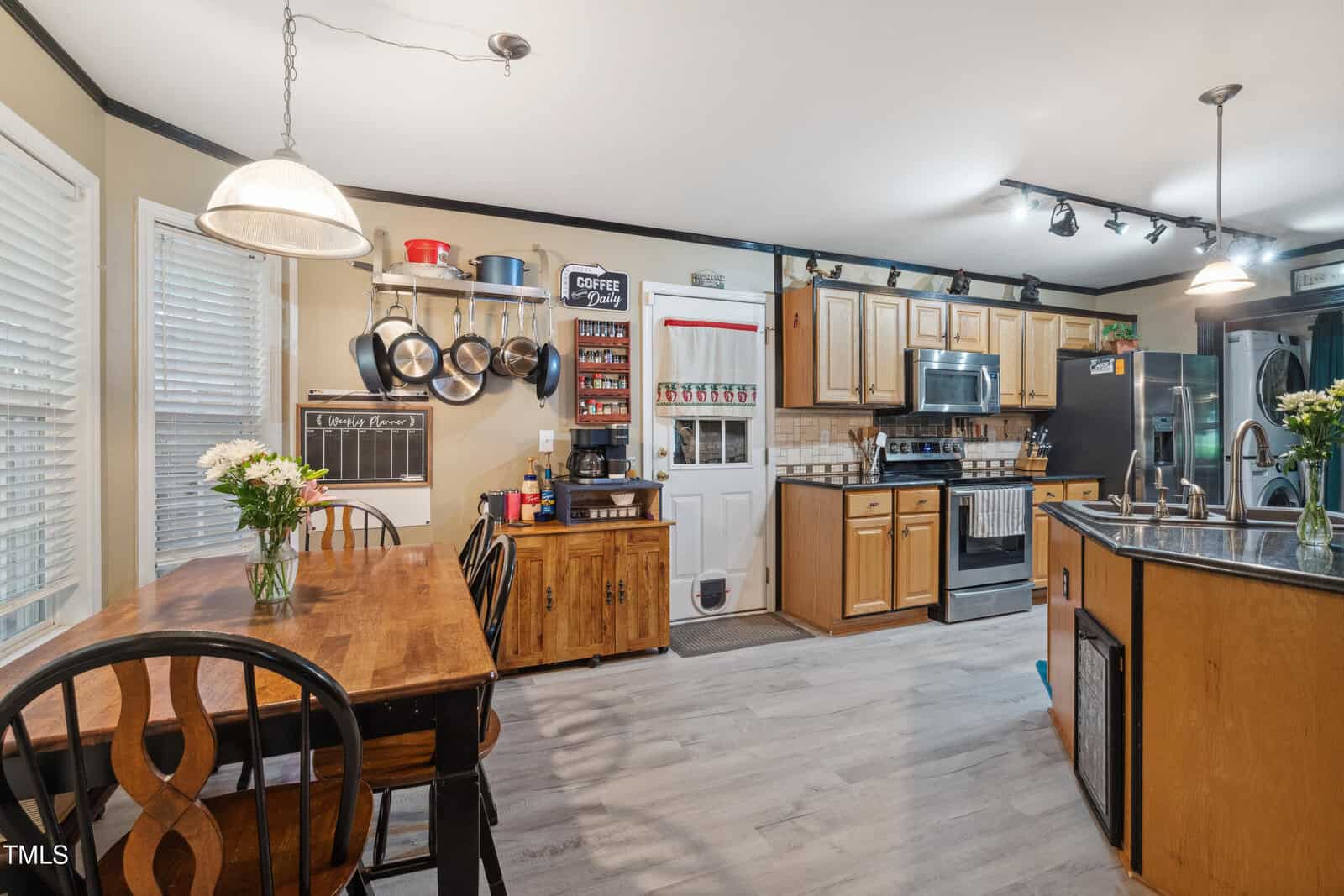 A kitchen with wood cabinets, stainless steel appliances, a dining table with chairs, flowers on the table, shelves with dishes, and a kitchen island with a sink. Light fixtures hang above the table and island.