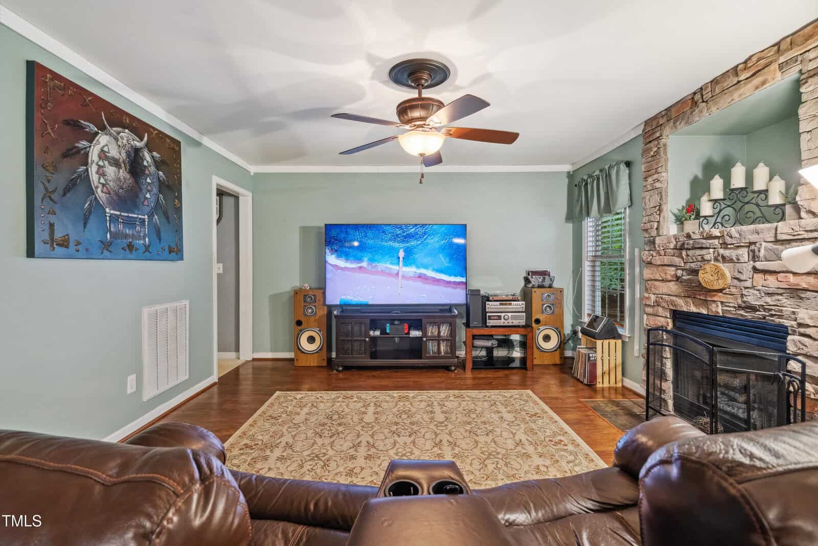 A living room with a brown recliner facing a large TV on a wooden stand, flanked by speakers. A stone fireplace is on the right, and a painting with a crab hangs on the left wall. Hardwood floors and a ceiling fan are visible.