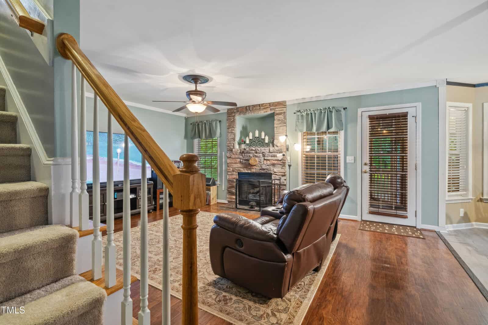 A cozy living room features a stone fireplace with a mantel, two brown leather recliners on a patterned rug, a ceiling fan, and wooden floors. Stairs with a wooden banister lead up on the left. Large windows bring in natural light.
