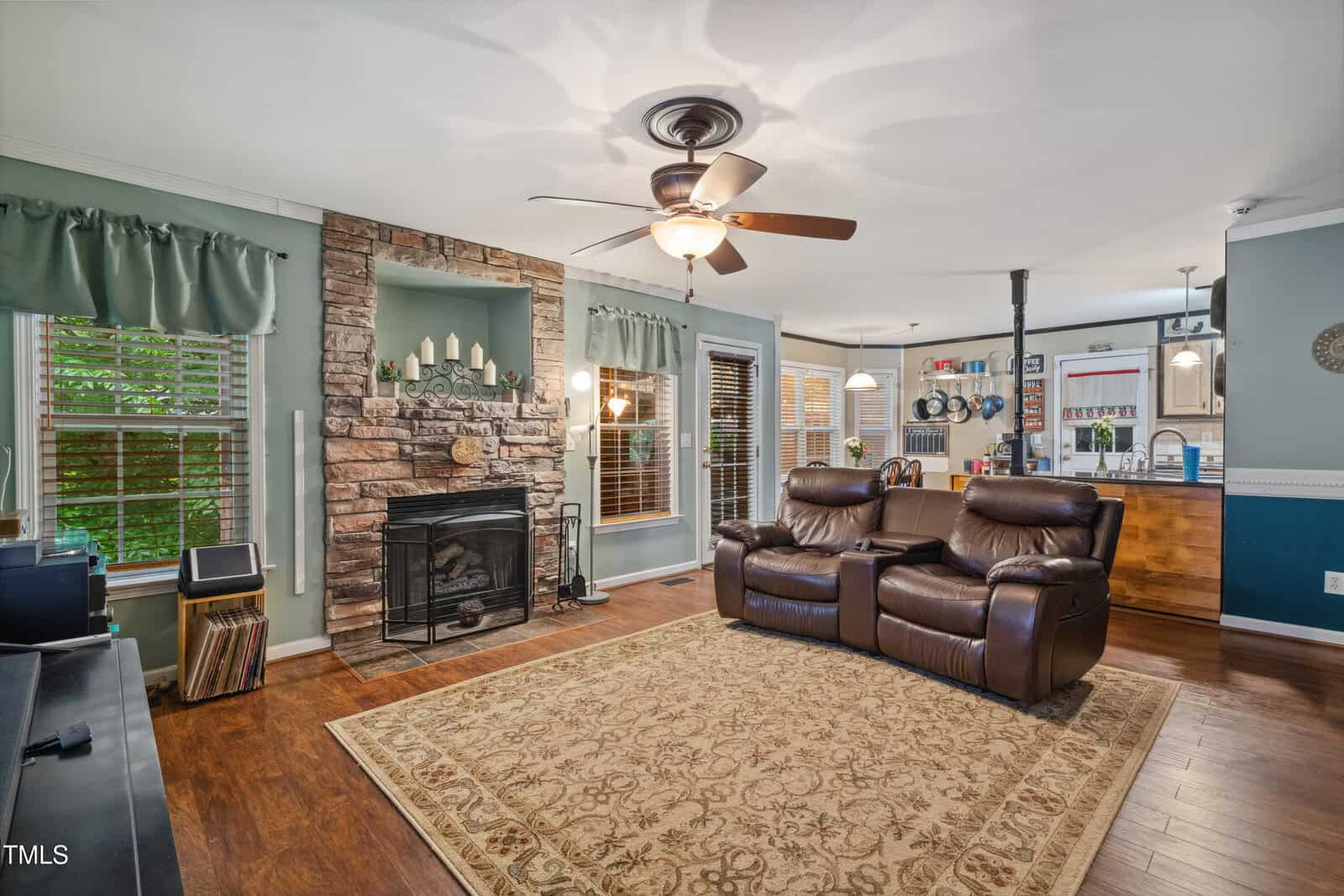 Living room with brown recliners, stone fireplace, ceiling fan, hardwood floors, and area rug. Kitchen and dining space visible in the background, with green curtains on windows and natural light coming in.