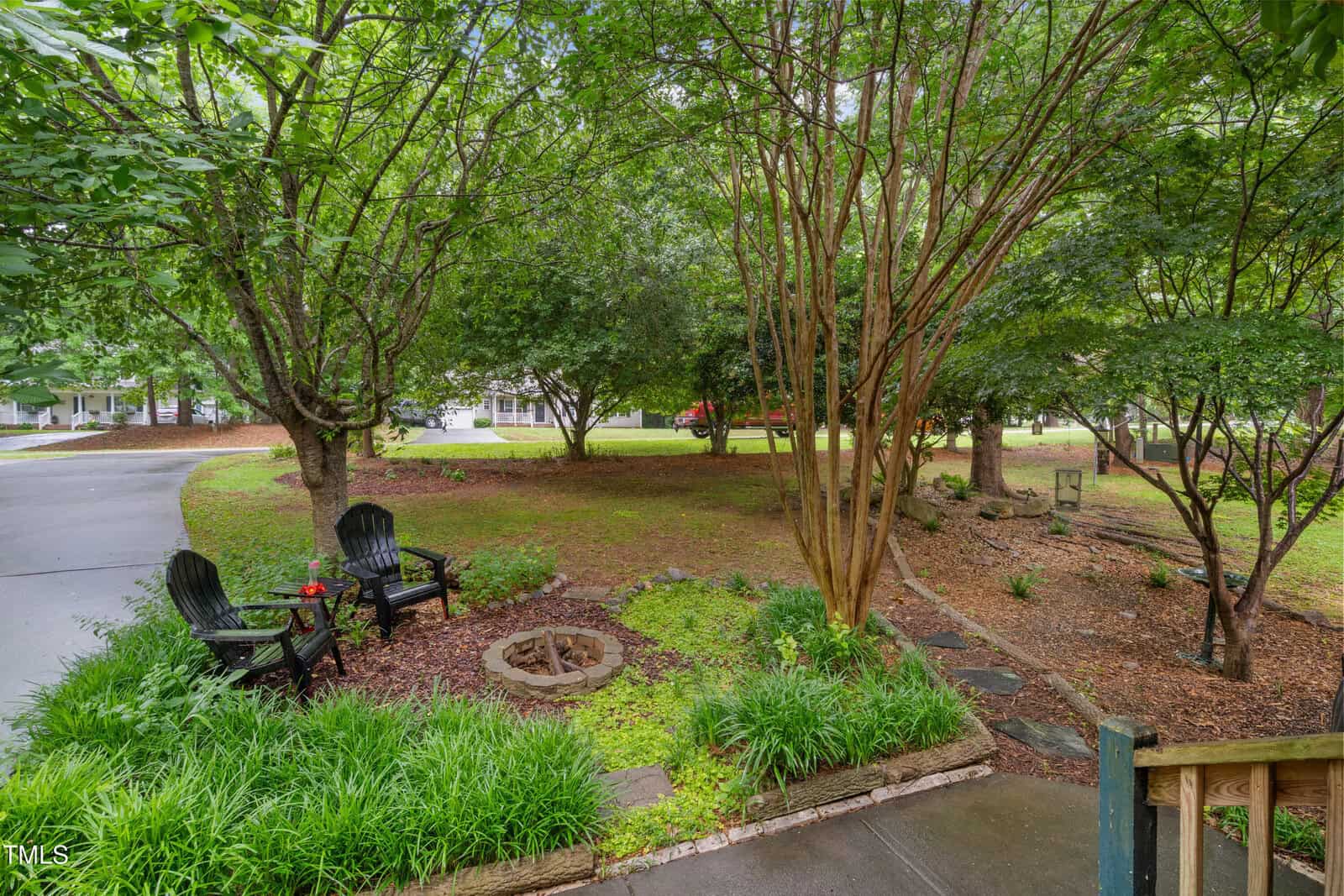 A lush, green yard with trees, a circular fire pit surrounded by two black chairs, and landscaped flower beds. A curved driveway and houses are visible in the background.