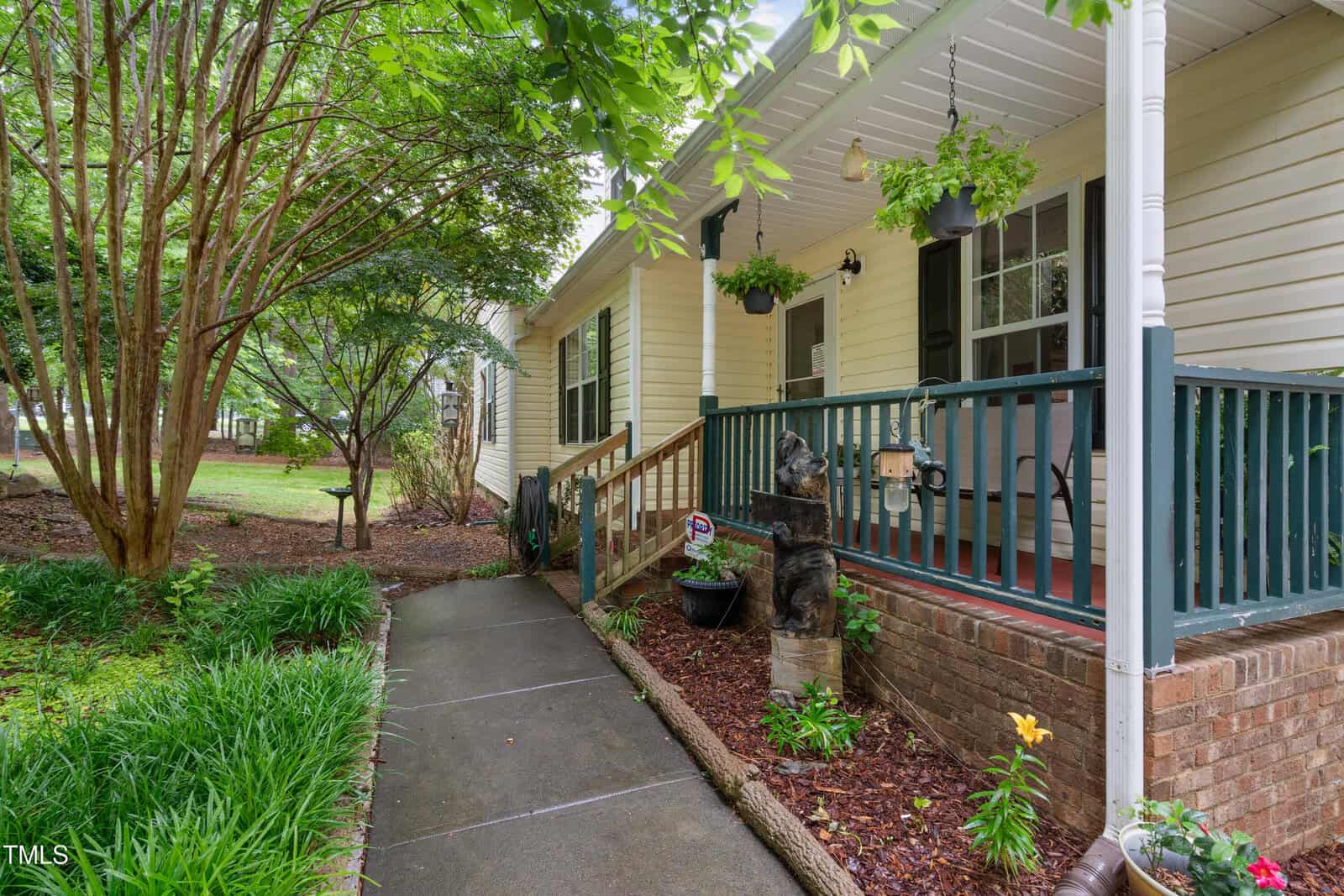 A yellow house with a covered front porch, green railing, hanging plants, a bear statue, and a sidewalk leading through a landscaped yard with trees and shrubs.