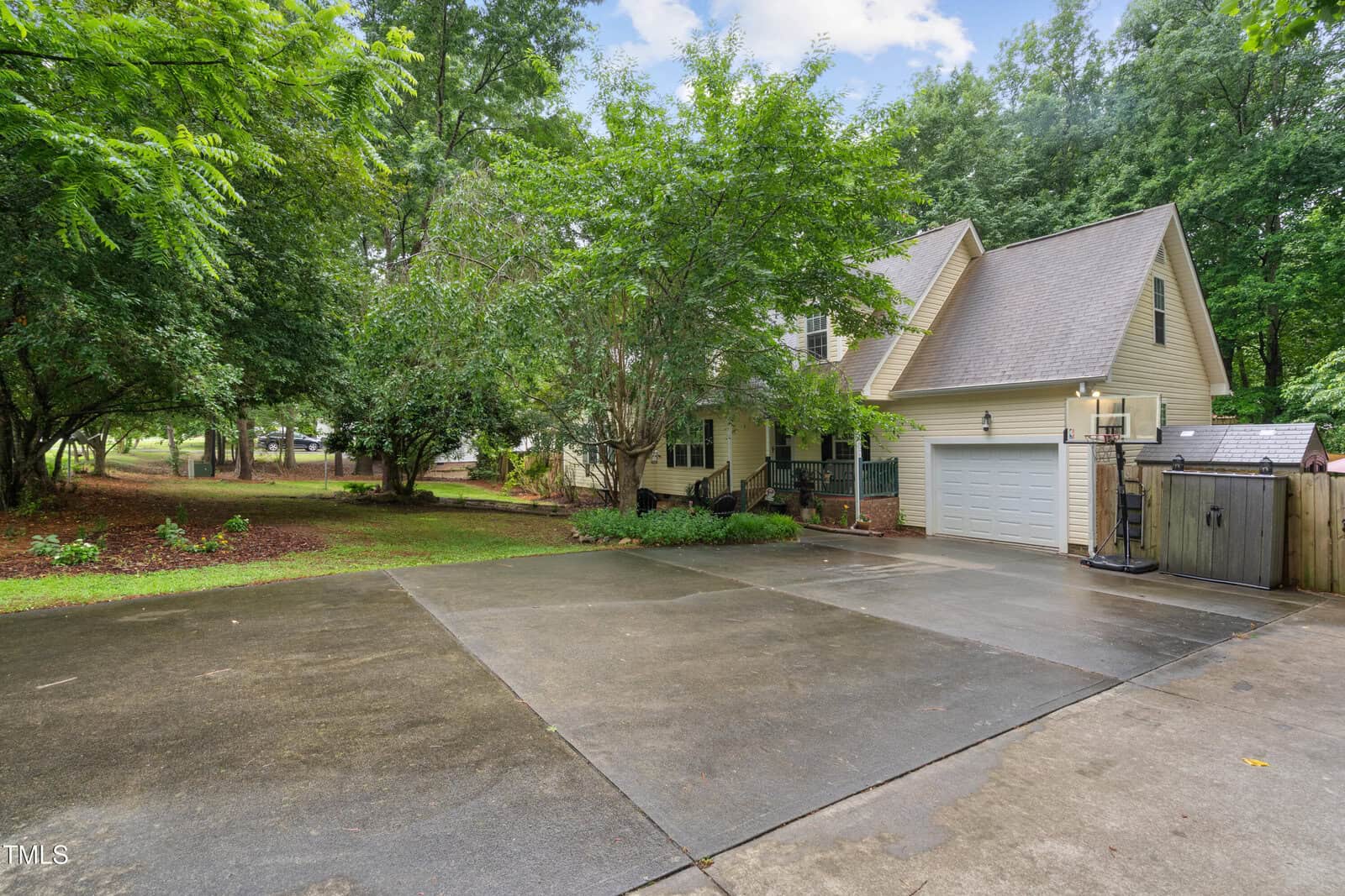 A two-story beige house with a gray roof, attached garage, and basketball hoop. Surrounded by trees and greenery, the home has a wide, wet driveway and a fenced backyard.
