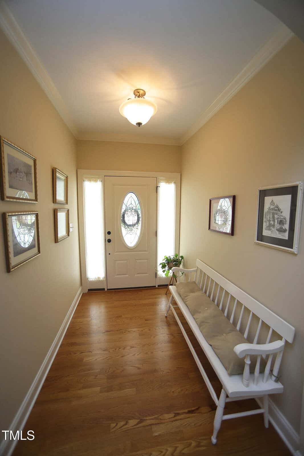 A bright hallway with light-colored walls, wood flooring, a white front door with glass panels, a cushioned white bench, a potted plant, and framed pictures hanging on both walls.