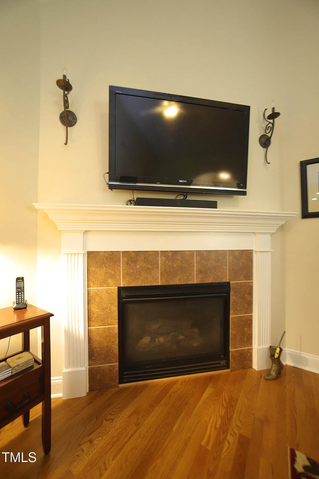 A flat-screen TV is mounted above a fireplace with a white mantel and brown tile surround. Two wall sconces flank the TV. Hardwood flooring and a small wooden side table with a remote are visible.
