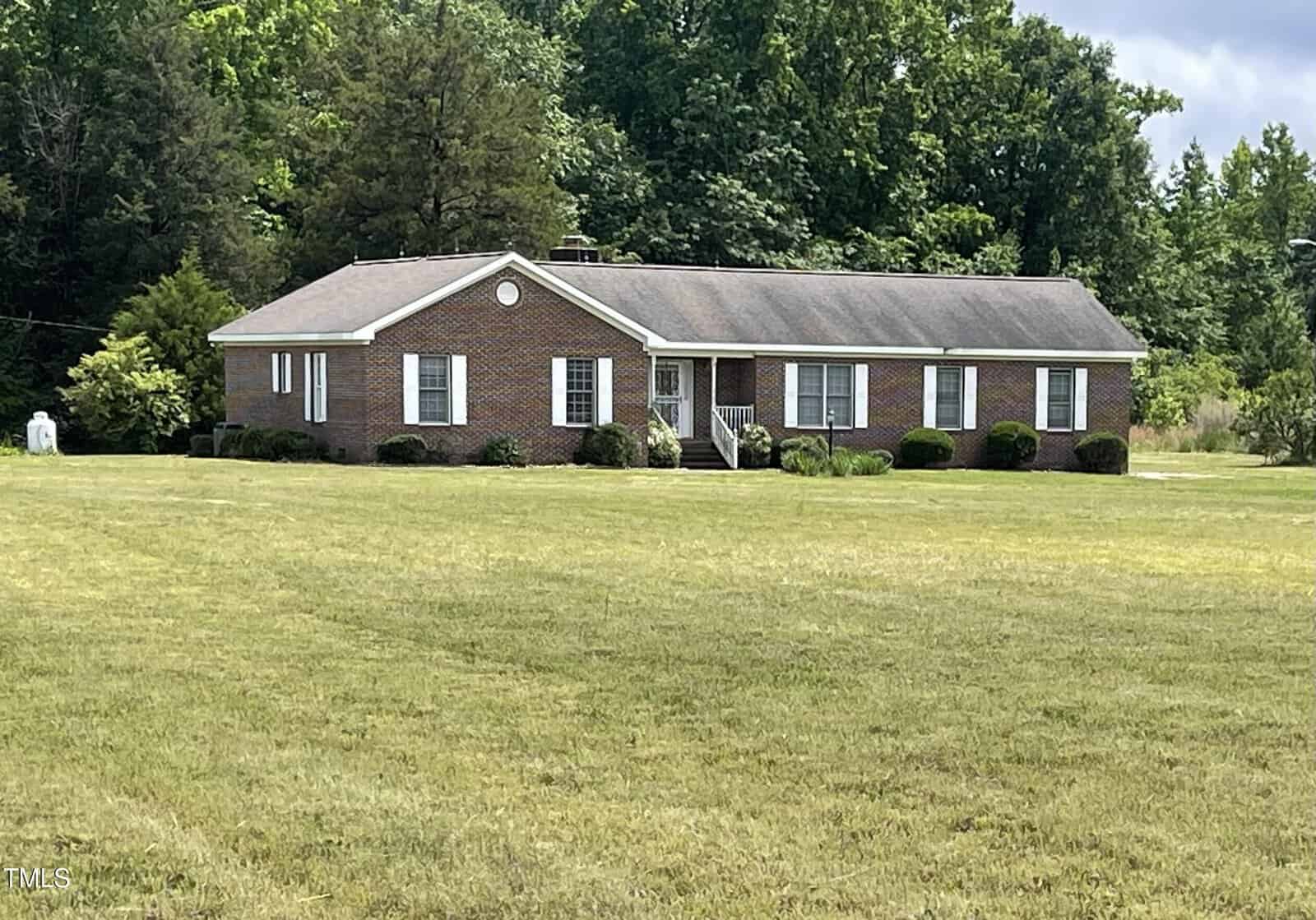 A single-story brick house with white trim and shutters sits on a large, well-maintained lawn, surrounded by trees and greenery in the background.