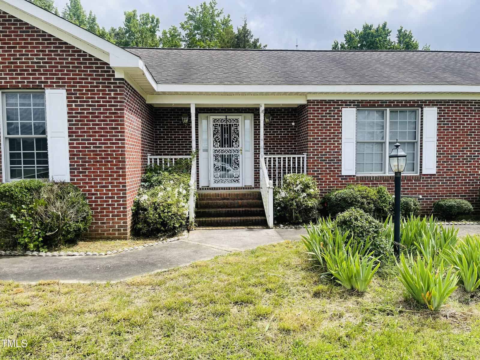 A single-story brick house with white trim at 65 Opie Frazier Road, Henderson. Steps lead to a metal screen door, with shrubs along the front, a lamp post, green plants in the yard, and a concrete walkway curving toward the porch.