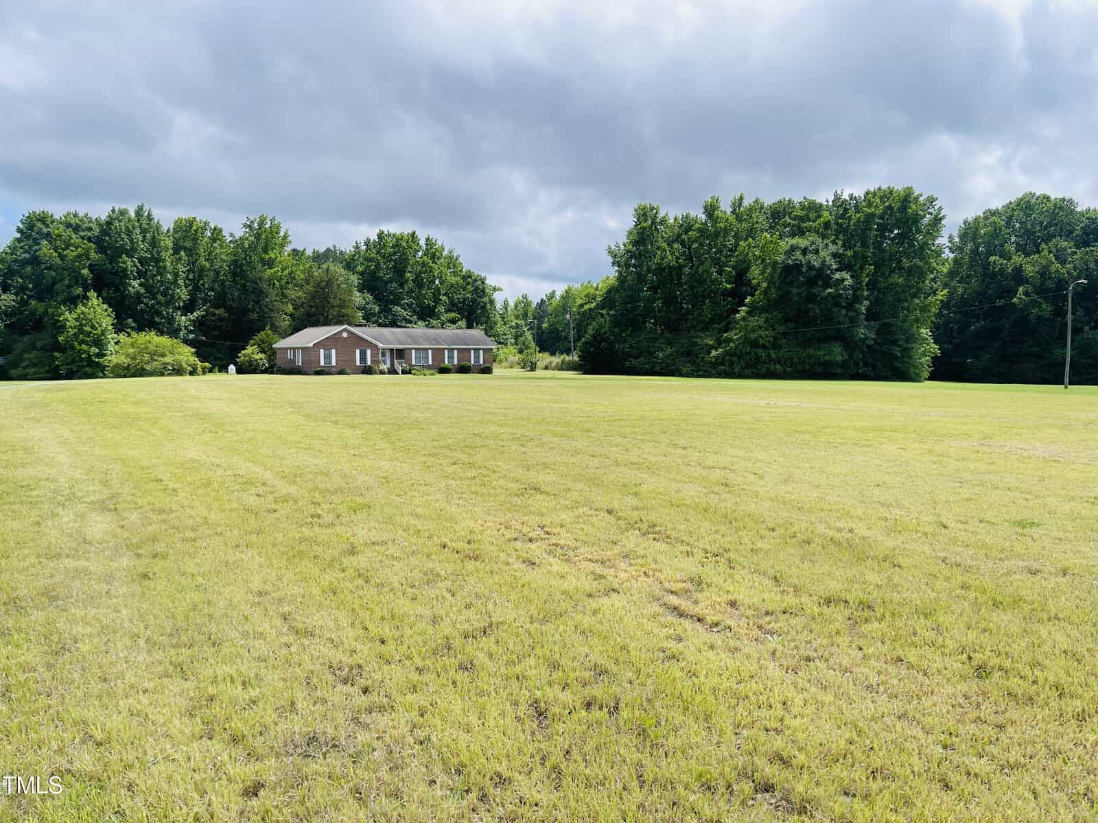 A single-story brick house is situated in the distance, surrounded by a large, open grassy field with dense green trees in the background under a cloudy sky.