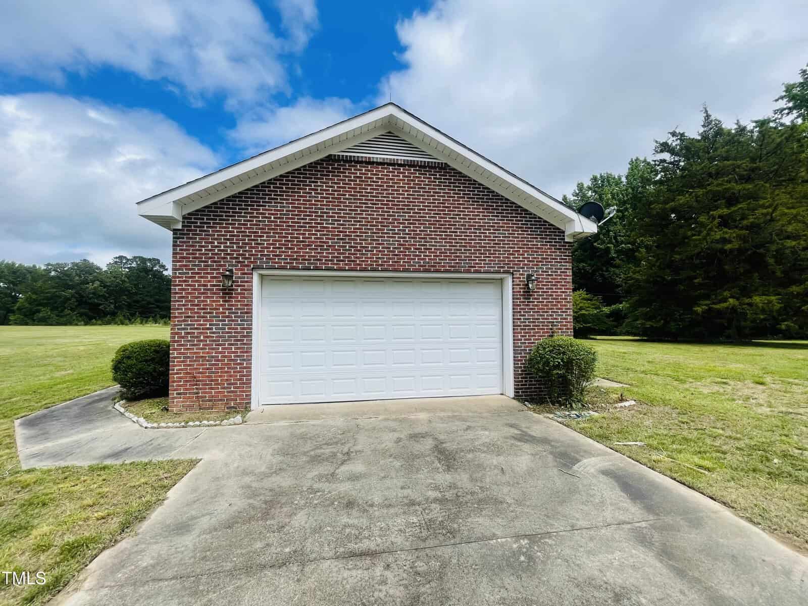 A two-car garage with a white door and brick exterior sits at the end of a concrete driveway, surrounded by trimmed bushes and grass, with trees and a cloudy sky in the background.