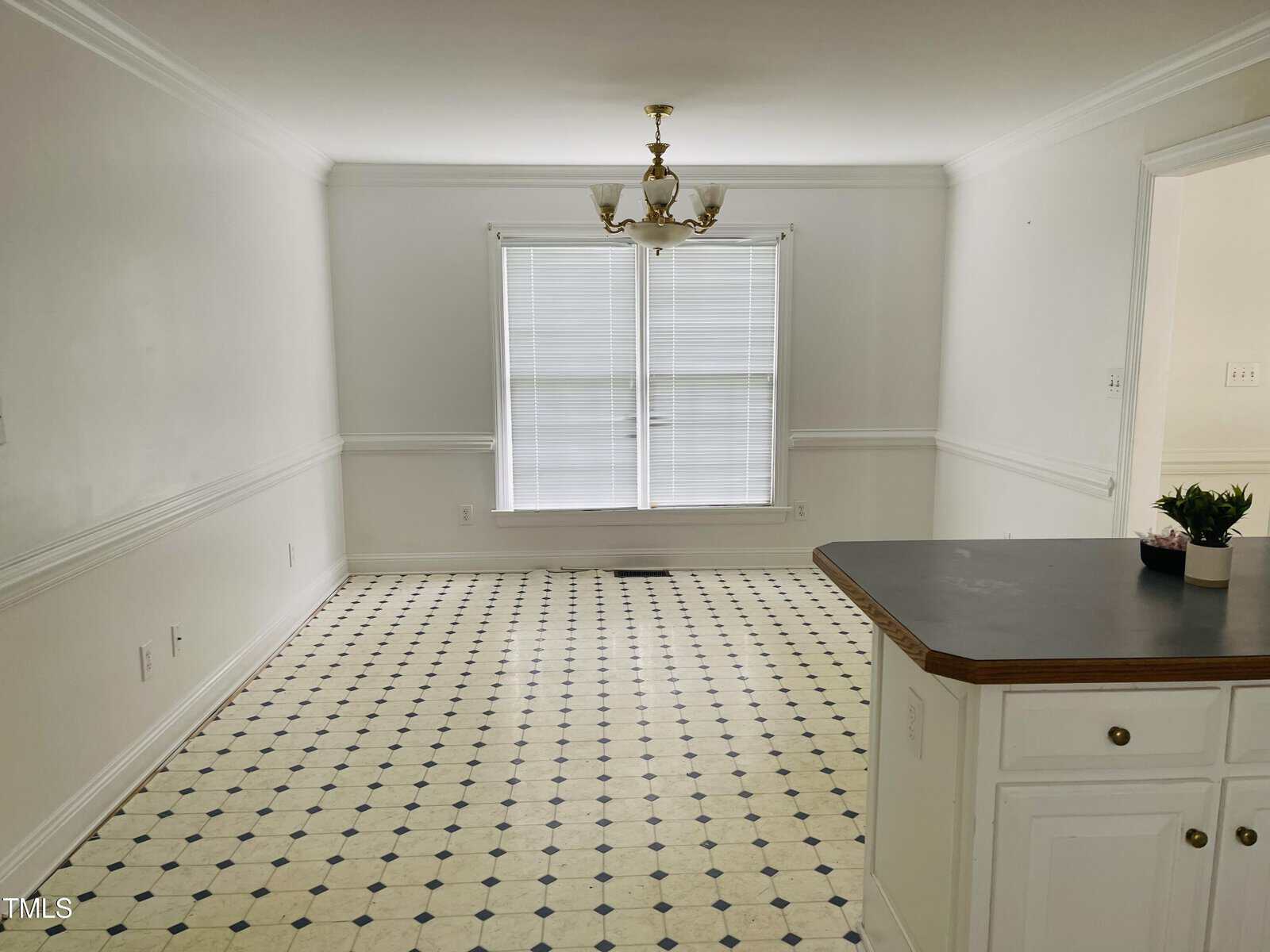 A bright, empty dining area with white walls and black-and-white tiled flooring. There is a chandelier in the center, two large windows with blinds, and part of a white kitchen counter with a small plant.
