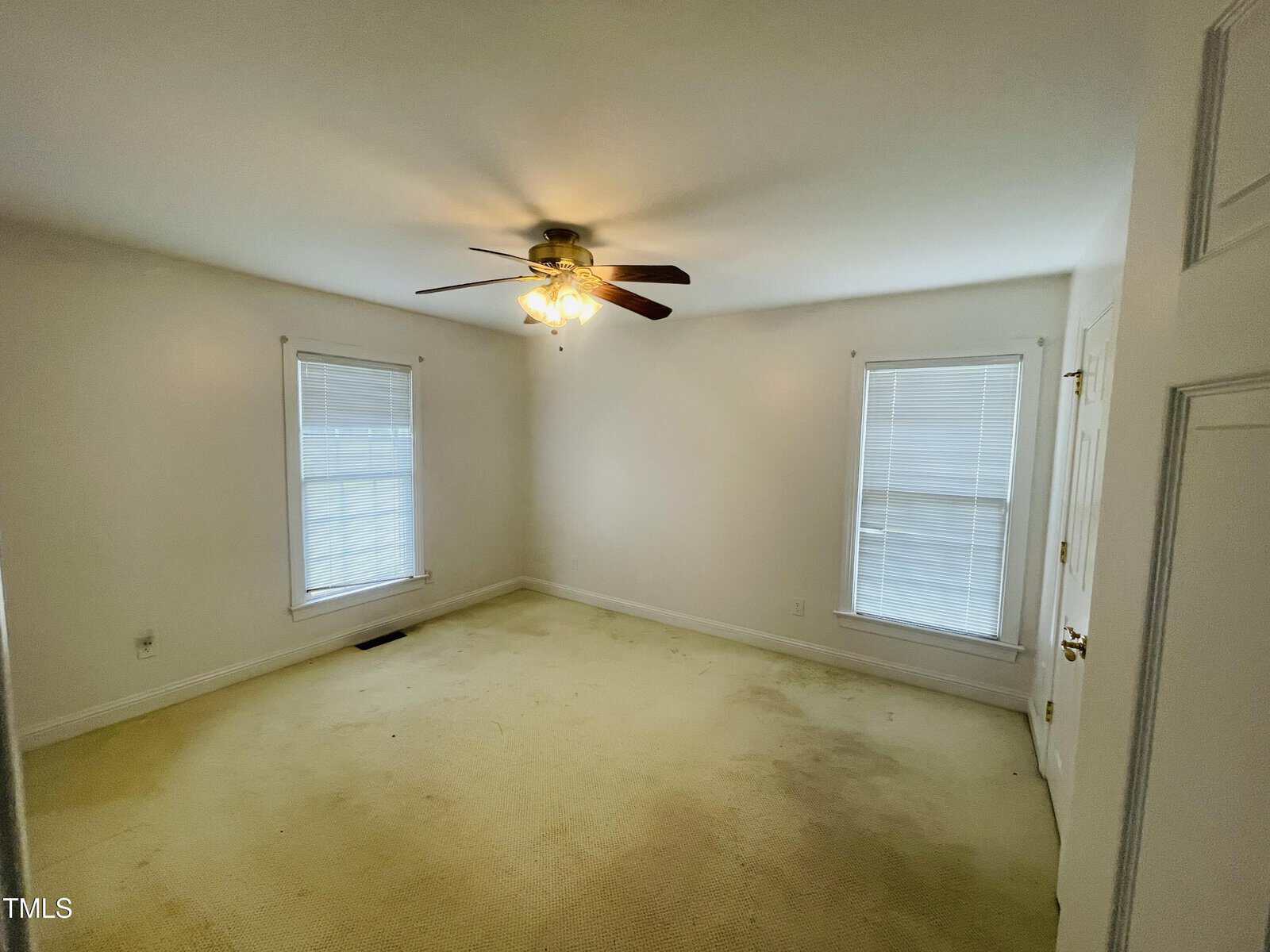 An empty bedroom with off-white walls, a beige carpet with visible stains, two windows with blinds, a white door, and a ceiling fan with lights turned on.