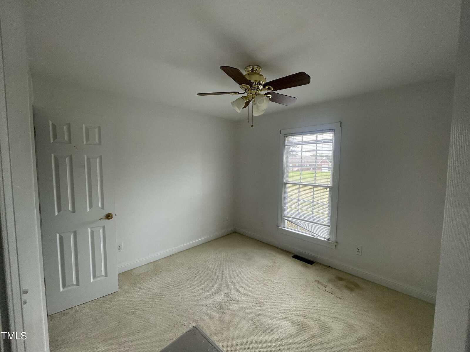 A small empty room with white walls, beige carpet, a ceiling fan with light, an open white door, and a window letting in natural light. There are visible stains on the carpet near the window.
