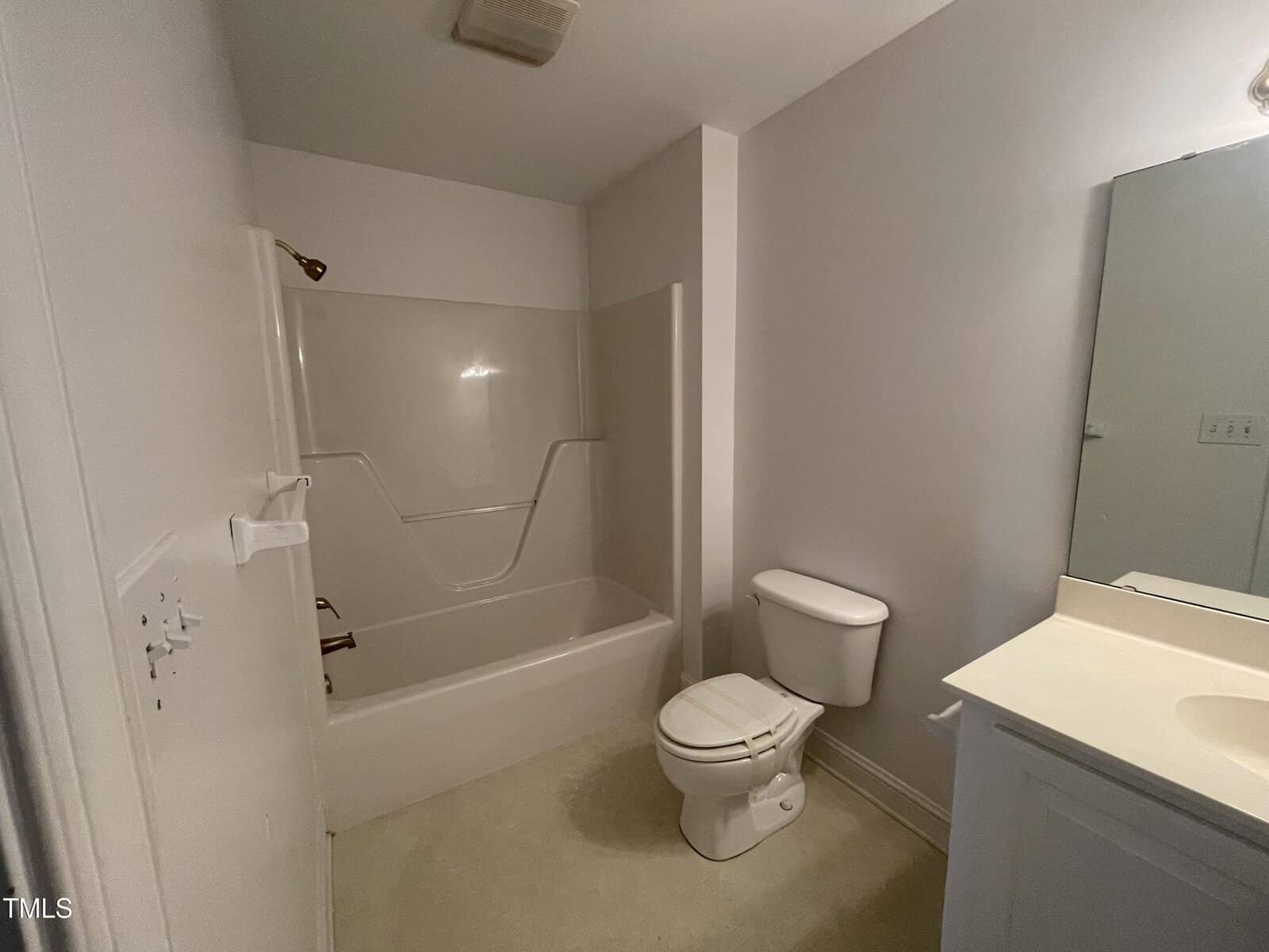 A simple bathroom with a white bathtub and shower combination, a white toilet, and a white sink vanity with a mirror above it. The walls and floor are light-colored, and the lighting is soft.