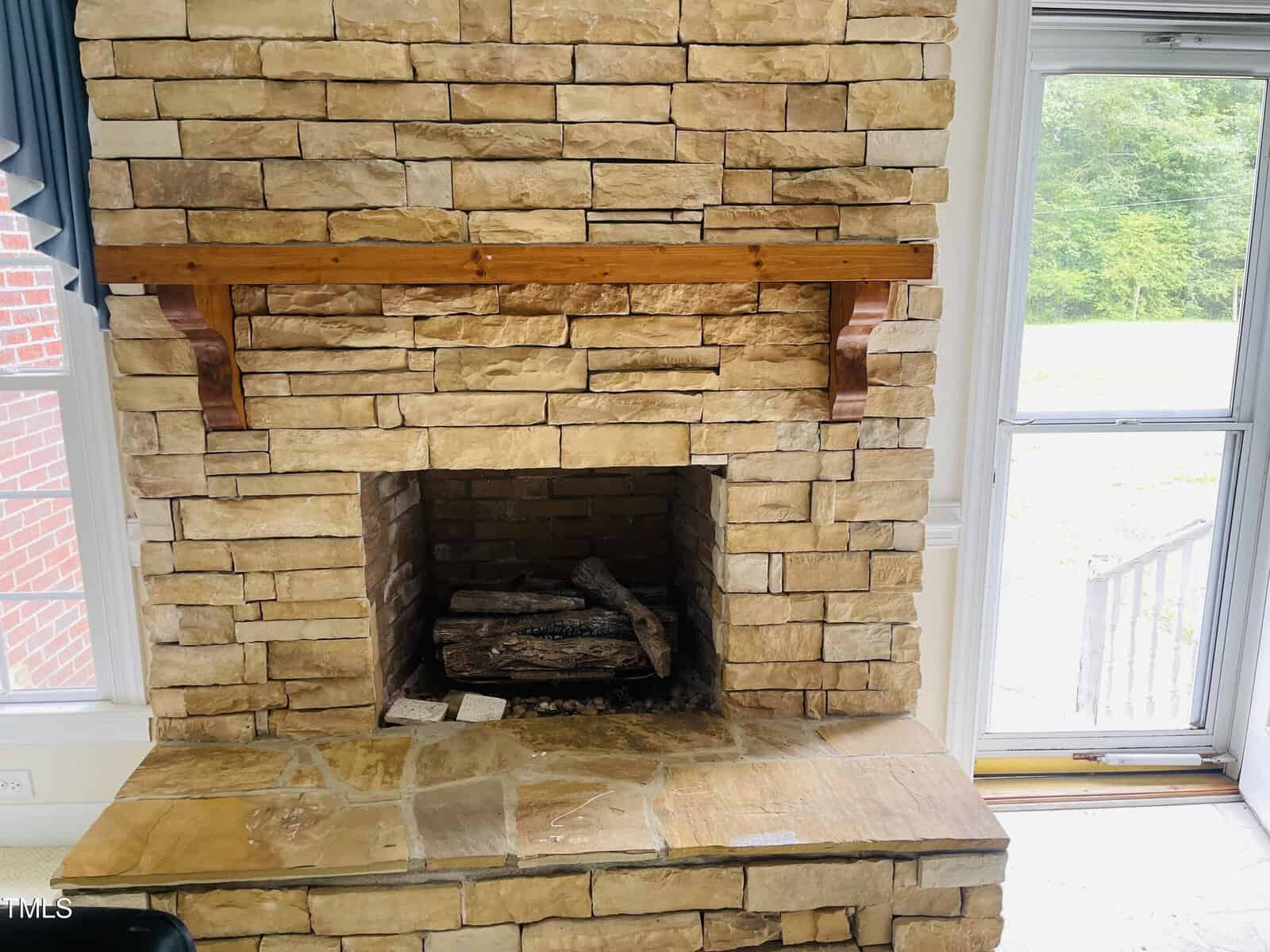 A stone fireplace with a wooden mantel, featuring stacked stone blocks and logs inside the firebox. A window is visible to the left and a glass door to the right, showing greenery outside.