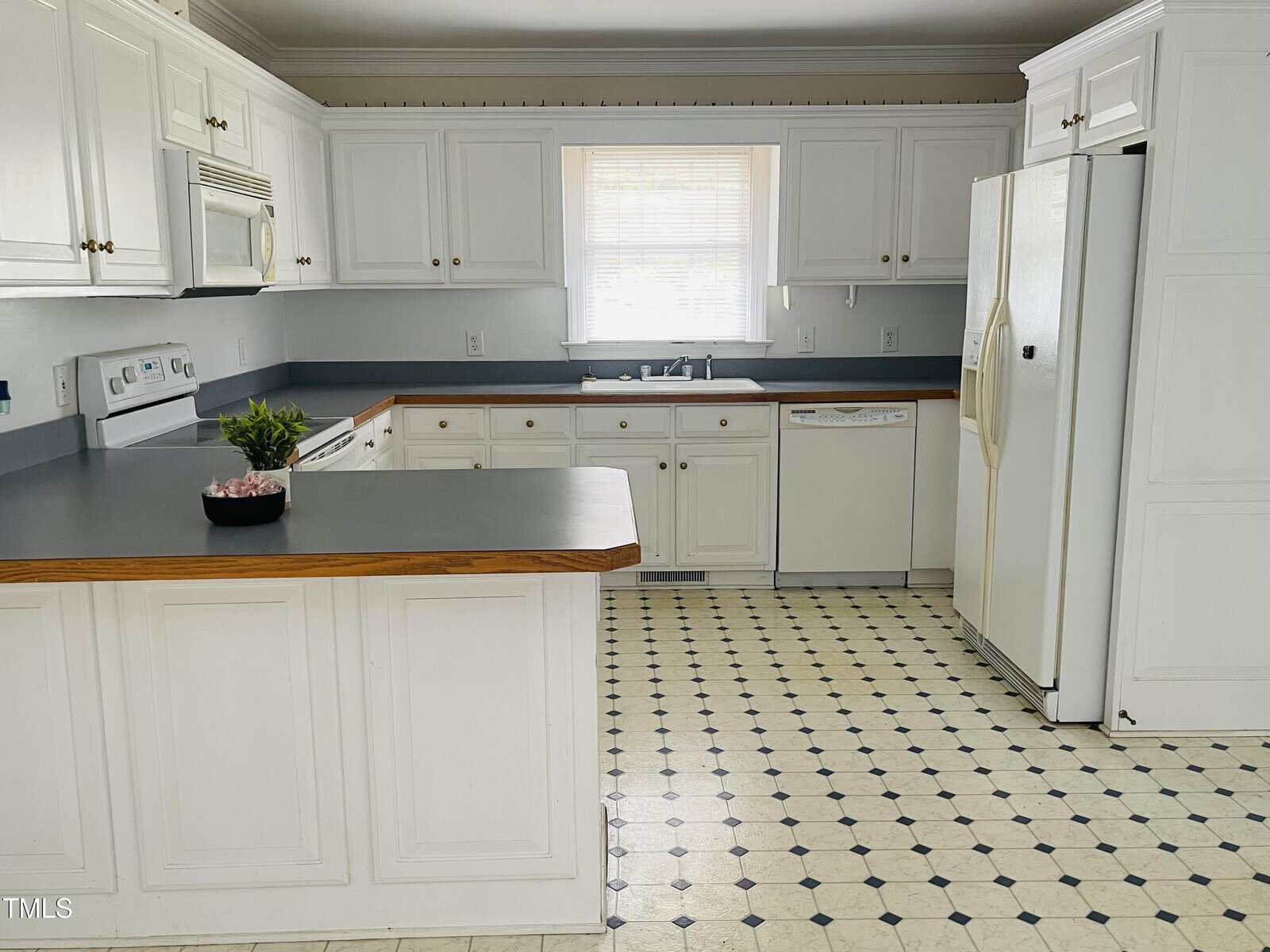A kitchen with white cabinets, white appliances, gray countertops, and a window above the sink. The floor has a white and black patterned linoleum, and there is a small green plant on the counter.