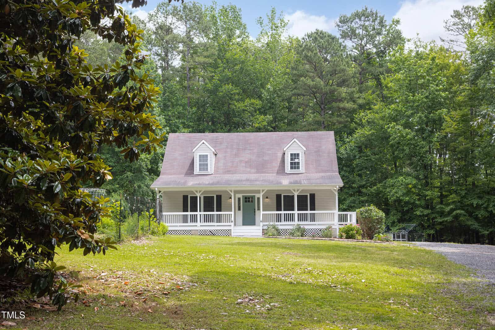 A white two-story house with a covered front porch and dormer windows sits on a grassy lawn, surrounded by trees and a gravel driveway, under a clear sky.