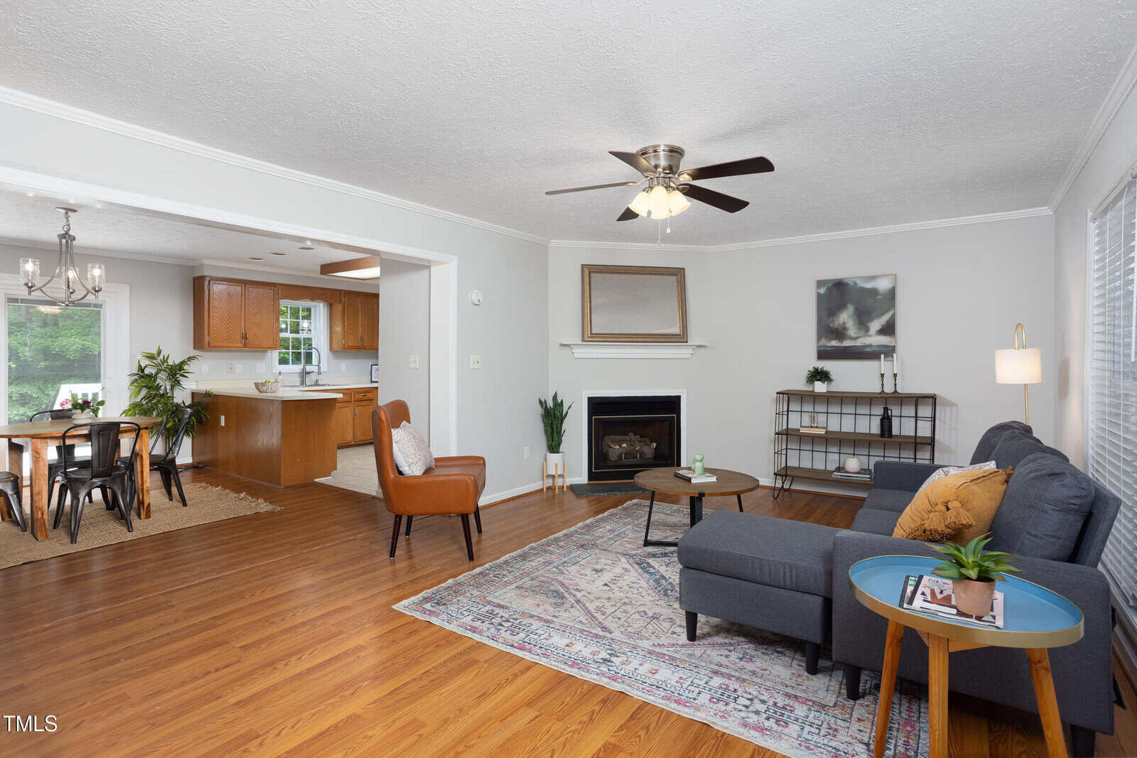 Bright living room with hardwood floors, gray sectional sofa, coffee table, and armchair. An open layout leads to a dining area and kitchen. Ceiling fan, fireplace, and large windows add light to the modern, cozy space.