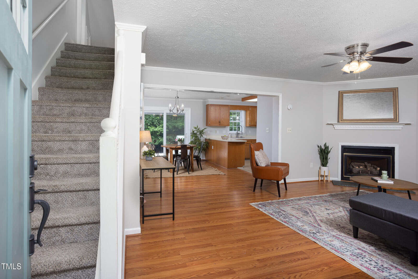 Open front door view of a living room with hardwood floors, a carpeted staircase on the left, fireplace with a mirror above it, and a dining area leading to a kitchen in the background.