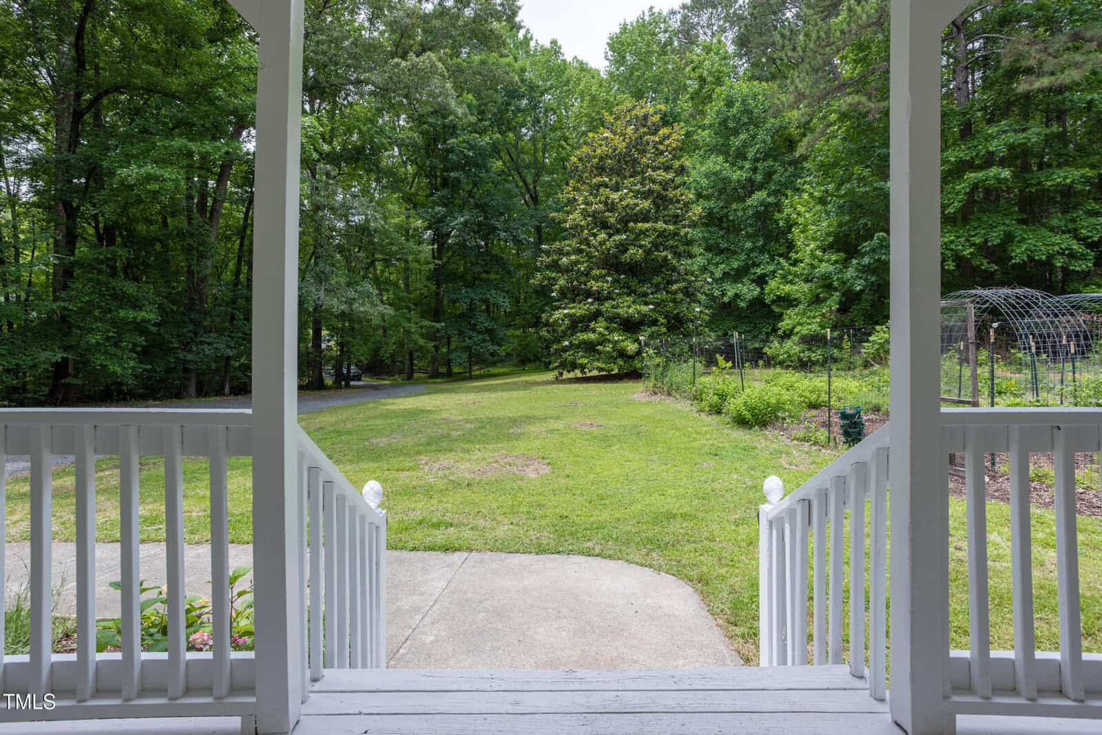 View from a covered porch looking out over a lawn with a concrete walkway, surrounded by trees and garden beds, on a bright, overcast day. White railings and steps frame the foreground.