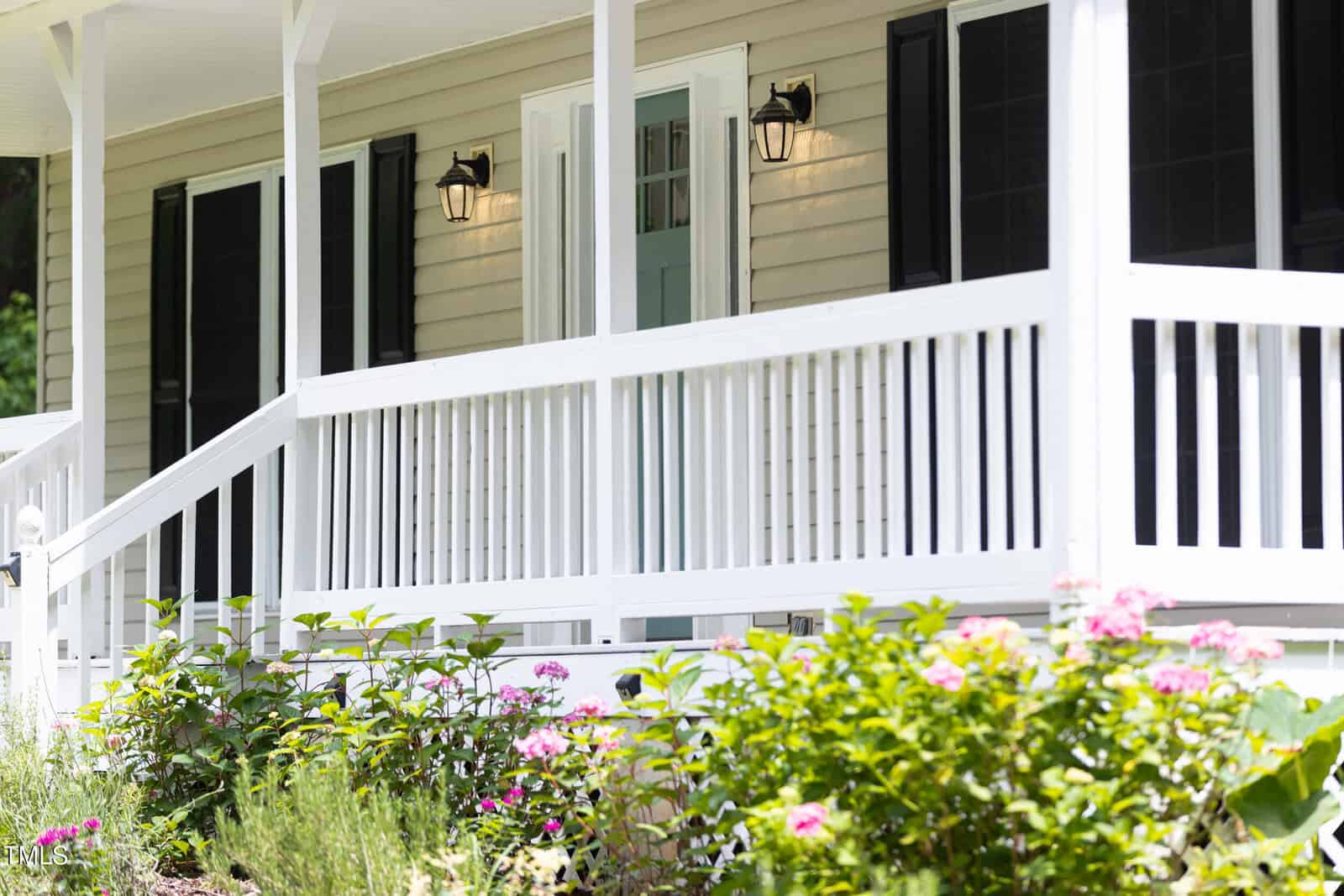 A white porch with railings and steps is attached to a beige house with black shutters. Two lantern-style lights flank a light blue door. Pink and green plants are visible in the foreground.