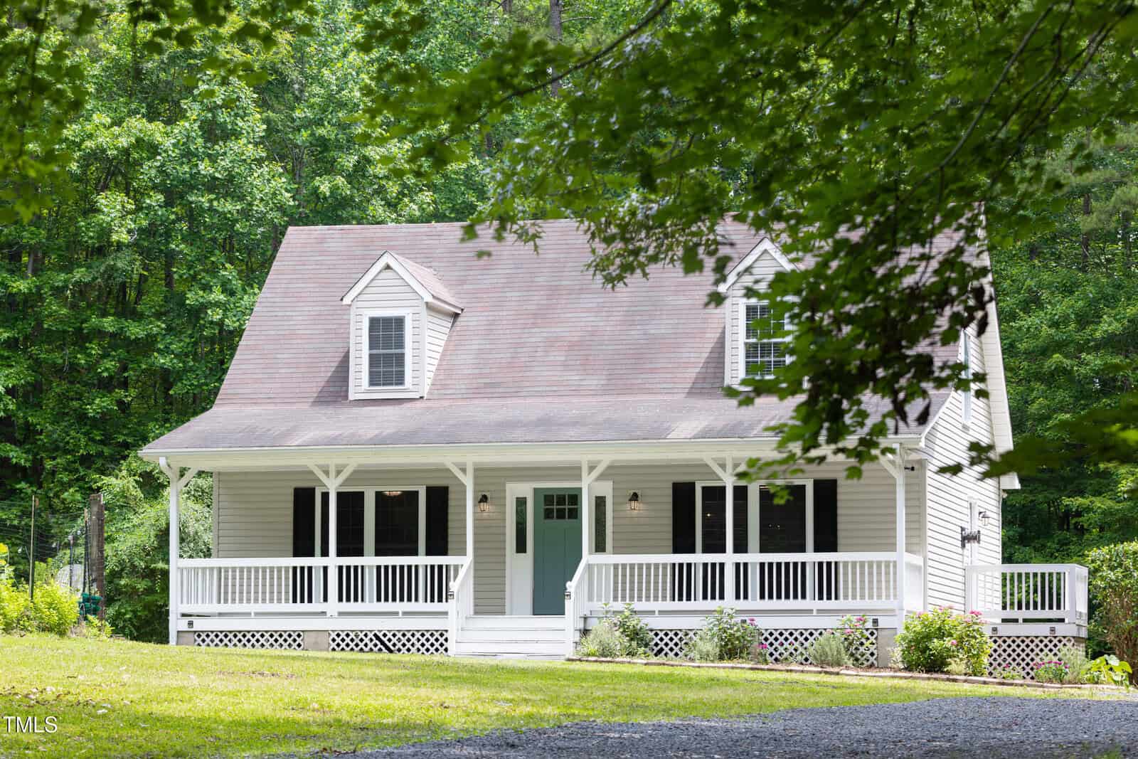 A light-colored, two-story house with a gable roof, front porch, white railing, and blue front door, surrounded by trees and greenery with a gravel driveway in front.