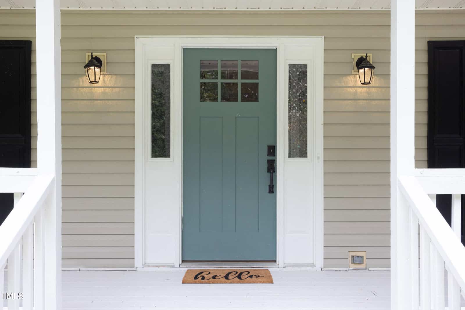 A light green front door with glass panes is centered between two white-framed windows on a beige house. Two black lantern-style lights are mounted on either side, and a doormat with "hello" is in front of the door.