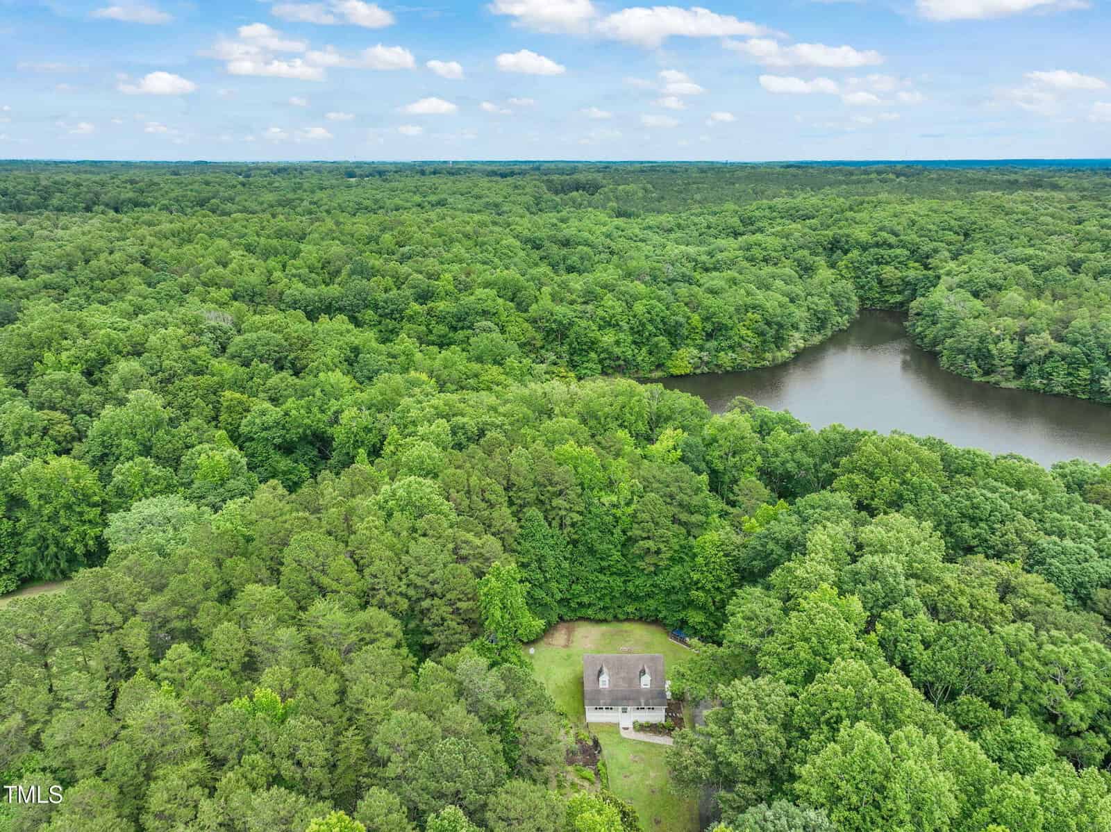 Aerial view of a house surrounded by dense green forest, with a small grassy yard and a lake visible nearby under a partly cloudy sky.
