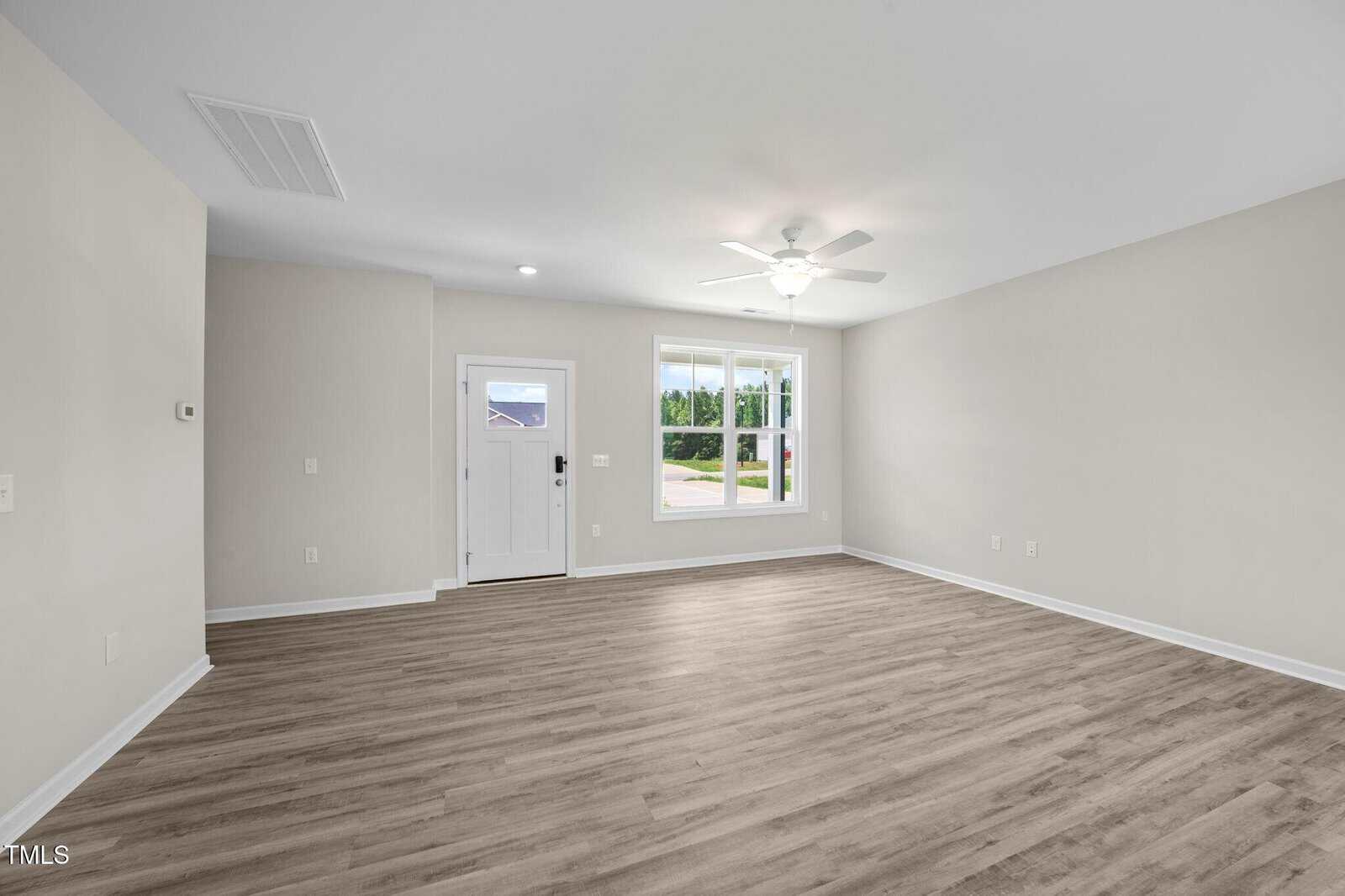 Empty room with light wood floors, beige walls, a white ceiling fan, and a large window. White front door with a window is on the left, and daylight is visible outside through the windows.