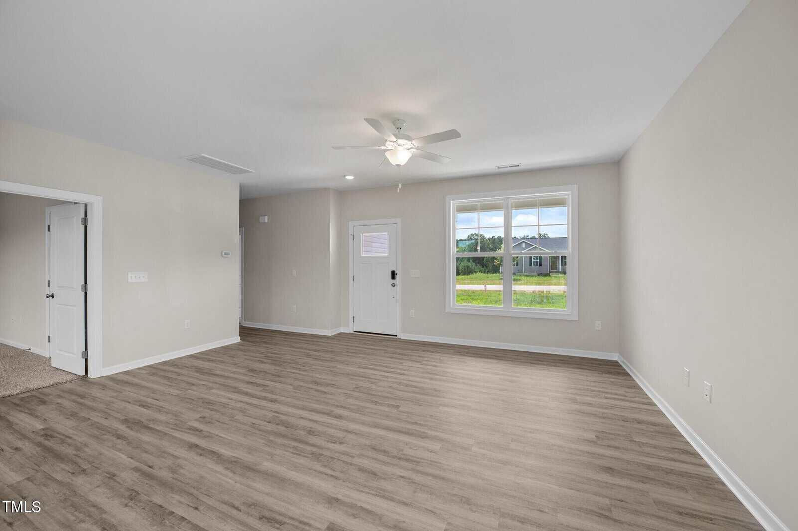 Empty living room with light gray walls, wood-look vinyl flooring, ceiling fan, single door with window, and large front window showing a lawn and house across the street. Double doors lead to another room.