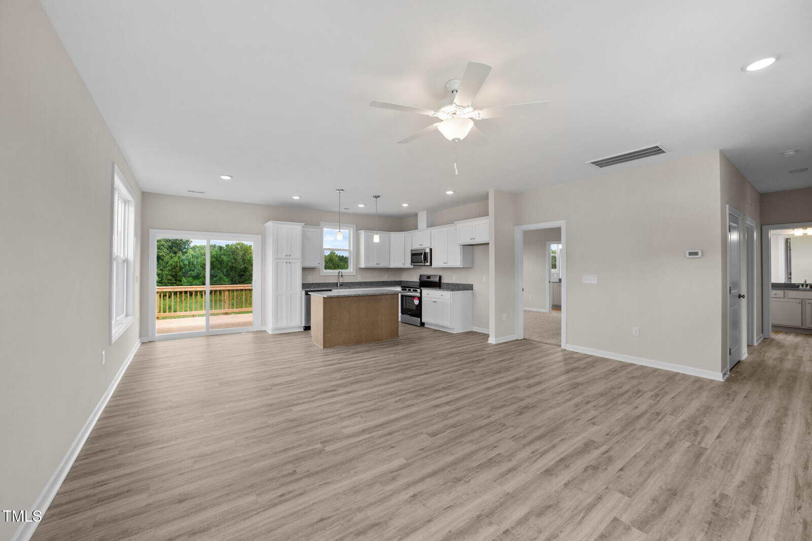 Open-concept kitchen and living area with light wood floors, white cabinets, stainless steel appliances, sliding glass doors to a deck, and adjacent bathroom and bedroom entrances. Ceiling fan and recessed lighting are visible.