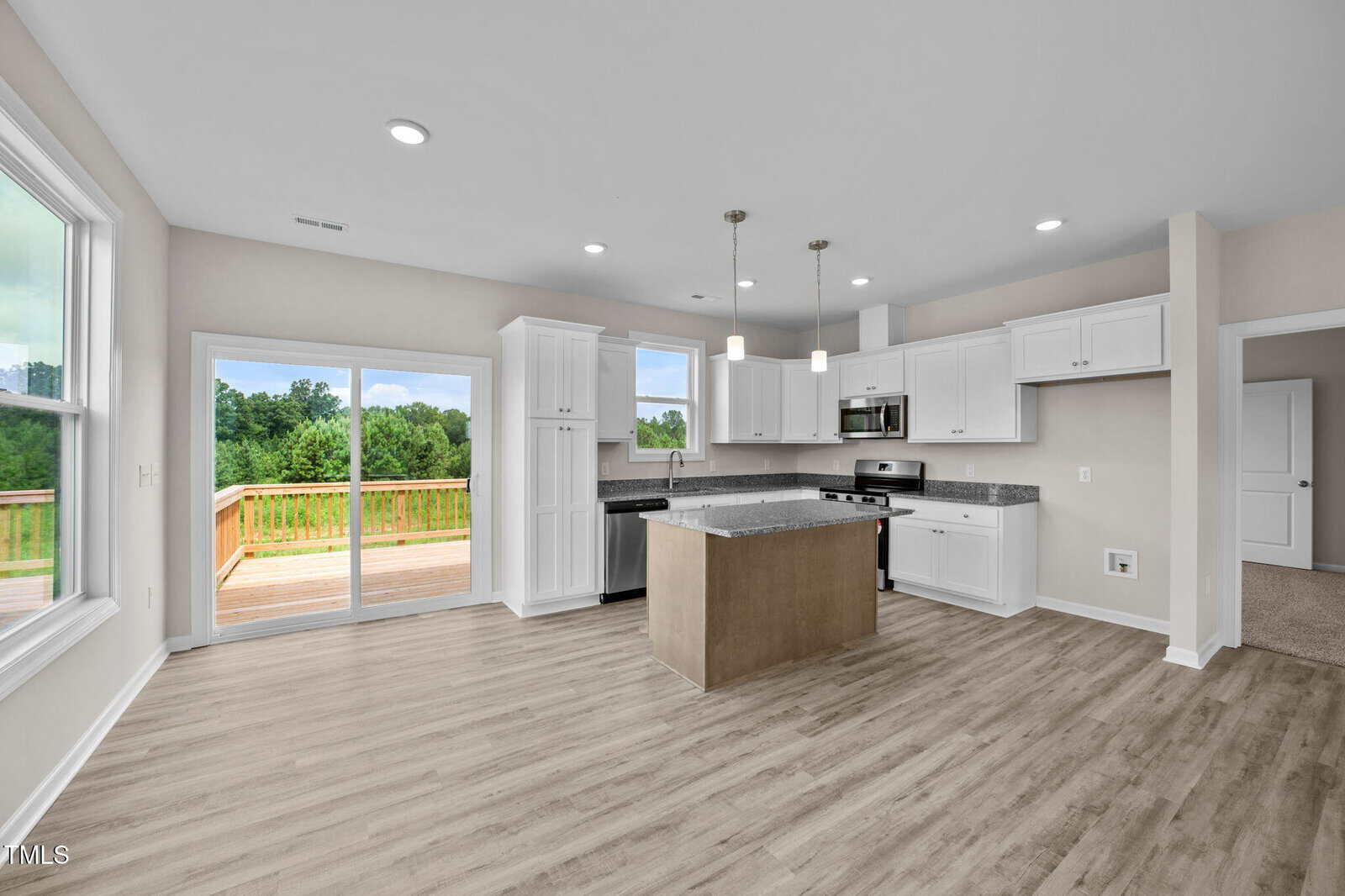 Modern kitchen with white cabinets, stainless steel appliances, a center island, and pendant lights. Large windows and sliding glass doors open to a wooden deck with a view of green trees. Light wood floors throughout.