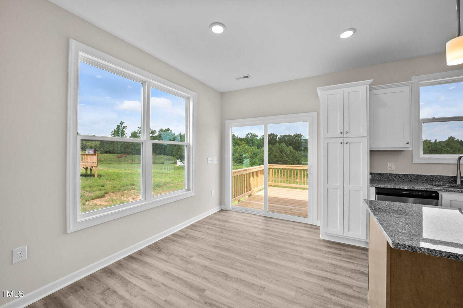 Bright dining area with large windows and sliding glass doors opening to a wooden deck, overlooking a green backyard. The space features light walls, wood flooring, and part of a modern kitchen.