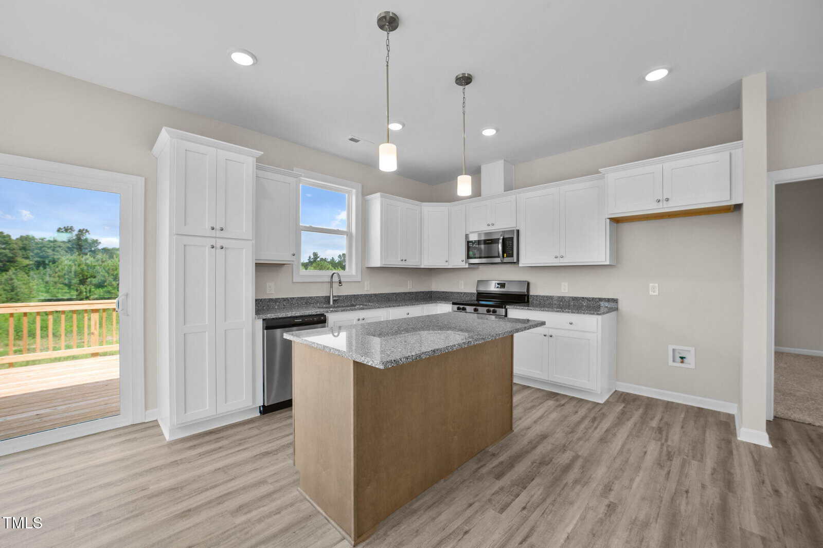 Modern kitchen with white cabinets, stainless steel appliances, a central wooden island with a granite countertop, light wood floors, pendant lights, and a sliding glass door leading to a deck with a view of greenery.