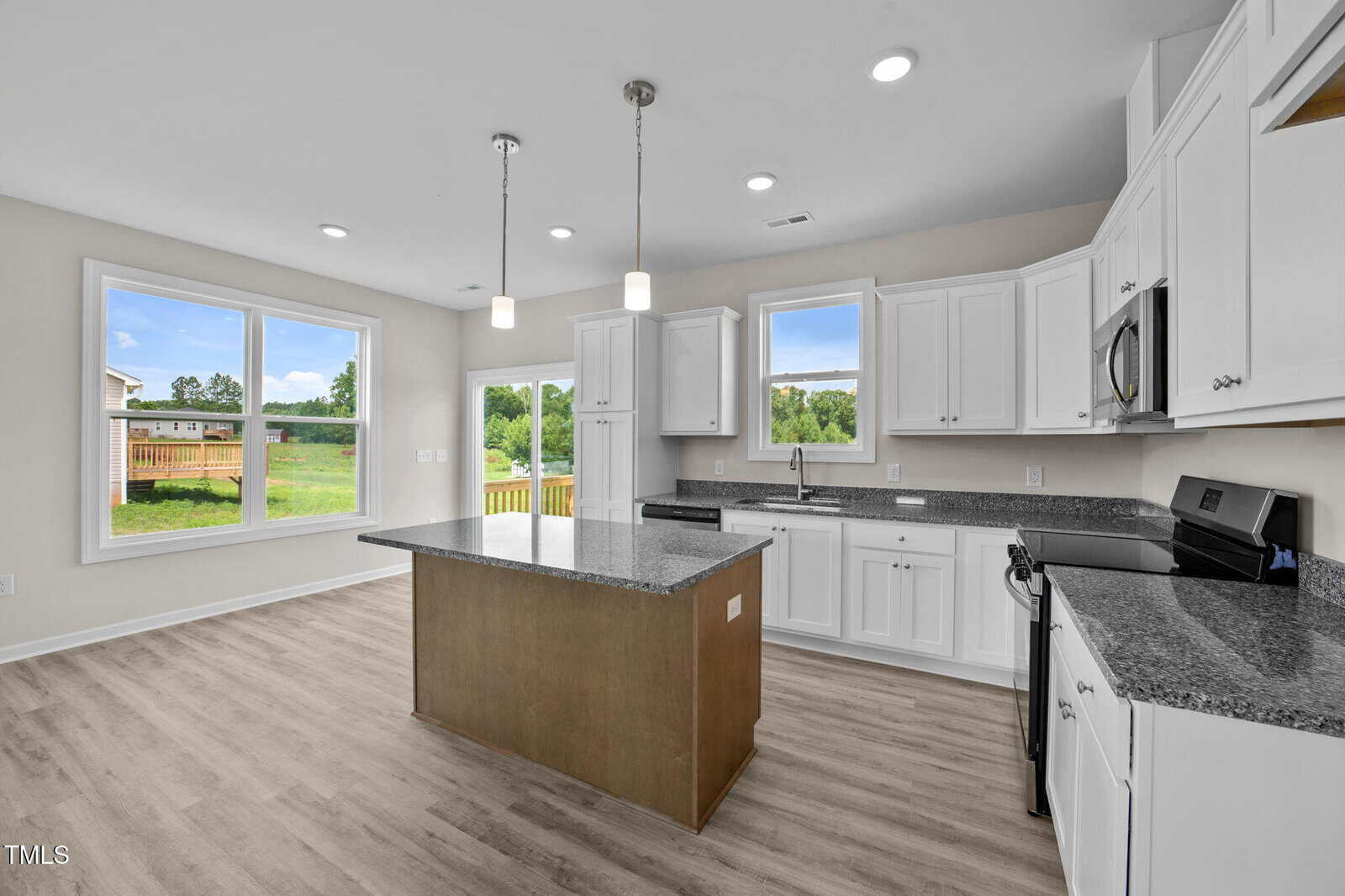 Modern kitchen with white cabinets, stainless steel appliances, granite countertops, a large island, and hanging pendant lights. Bright space with large windows overlooking a grassy backyard and deck. Light wood flooring throughout.