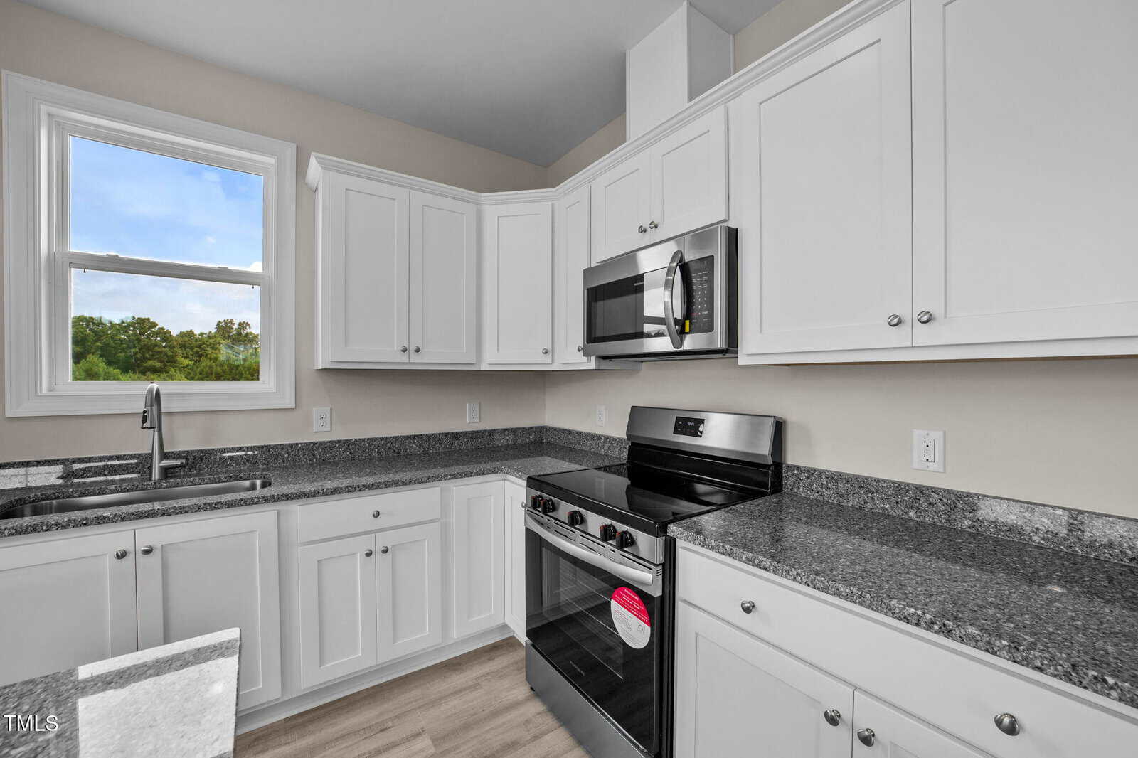 Modern kitchen with white cabinets, stainless steel appliances including a stove and microwave, granite countertops, a double sink beneath a window, and light wood flooring.
