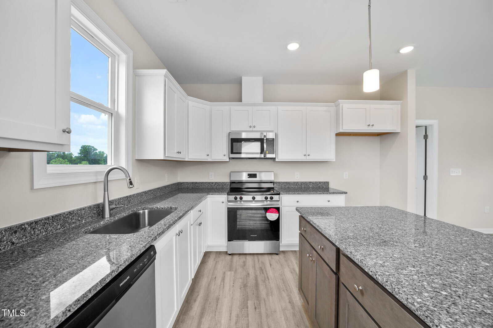 Modern kitchen with white cabinets, granite countertops, stainless steel appliances, a center island with dark wood base, a double sink under a window, and light wood flooring. Bright, clean, and spacious.