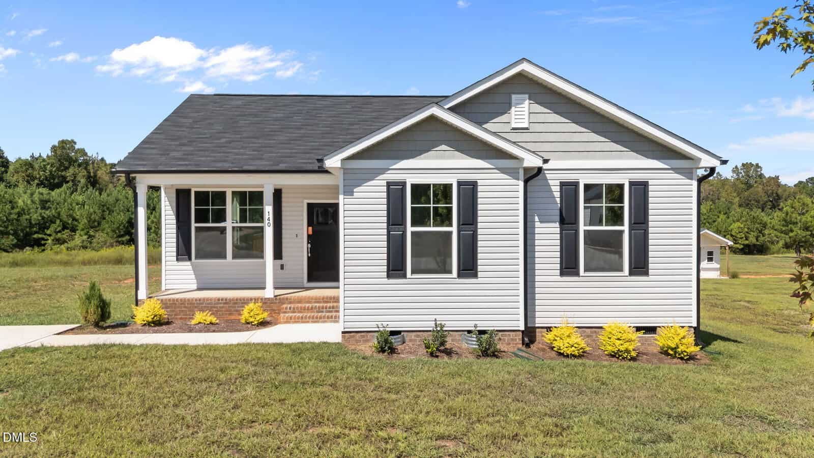 Charming single-story white house at 140 Brookhaven Drive, Spring Hope, with black shutters, a covered front porch, and a well-kept lawn. Small shrubs and yellow plants line the front, framed by trees under a clear blue sky.