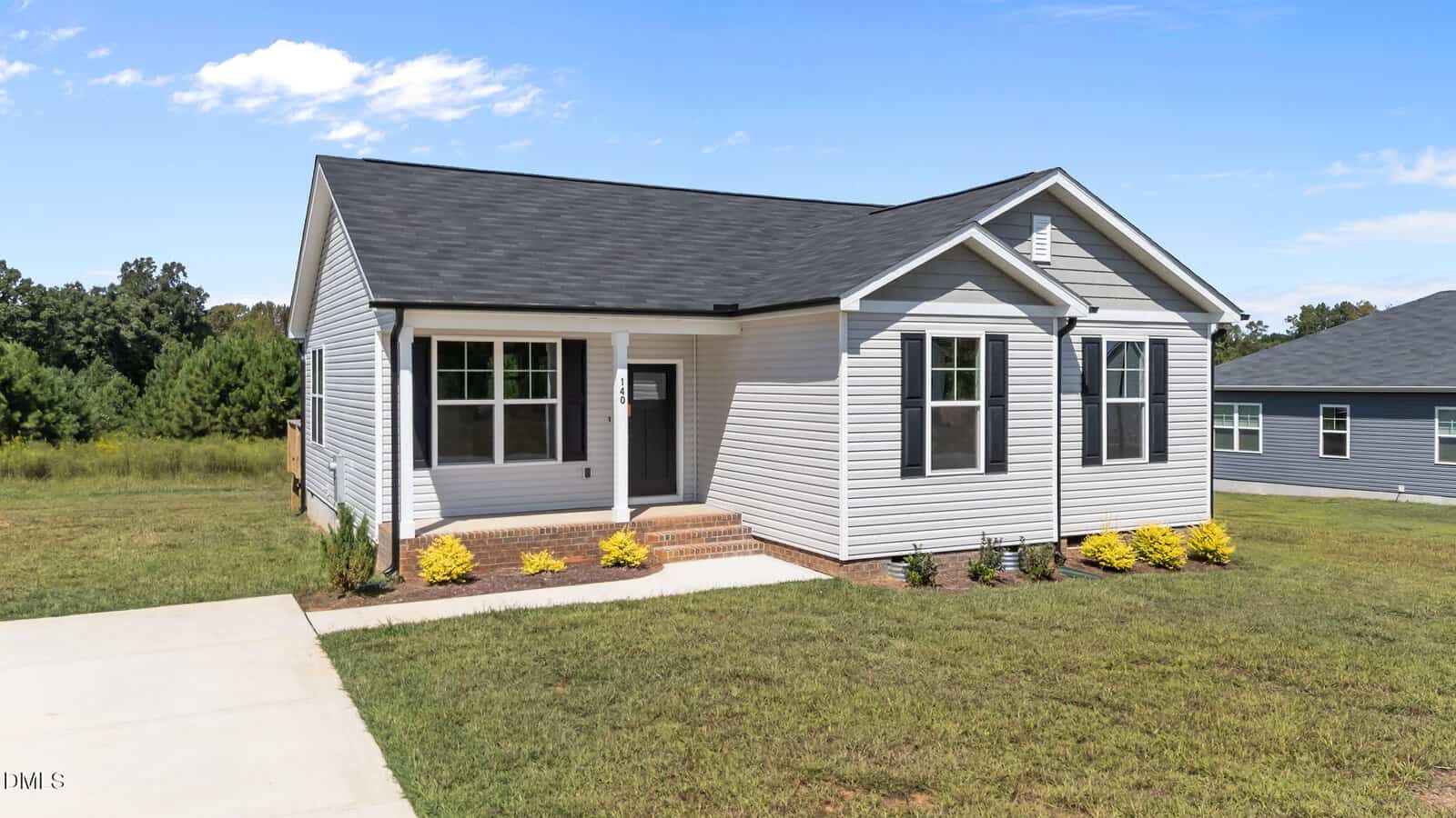 A single-story, light gray house with black roof shingles, white trim, and a small front porch with brick steps. The yard is grassy with neat landscaping and a concrete driveway. Trees and a blue sky are in the background.