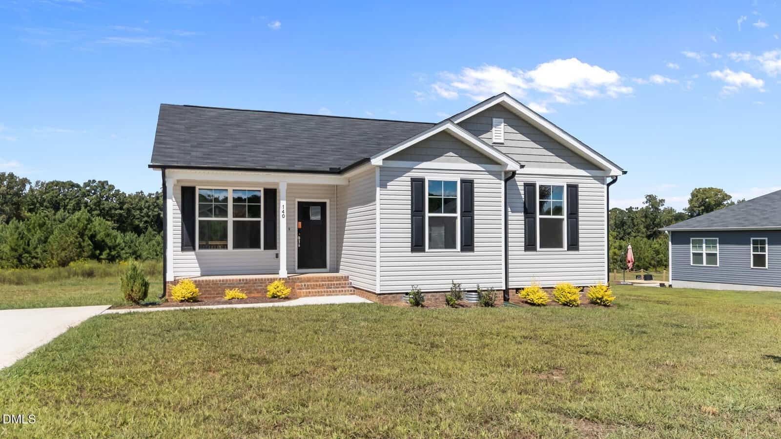A small, modern single-story house with gray siding, black roof, and white trim, featuring a front porch, steps, manicured shrubs, and a spacious green lawn under a bright blue sky.