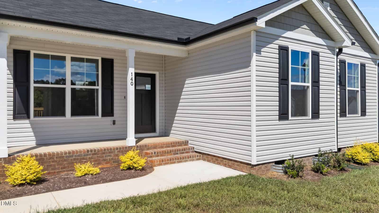 Single-story house with light-colored siding, dark shutters, large front window, and a black roof. Brick steps lead to a covered entryway. Small shrubs and plants line the walkway and foundation.