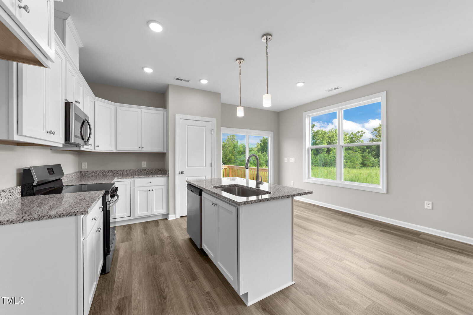 Modern kitchen with white cabinets, granite countertops, stainless steel appliances, a center island with pendant lights, and wood-look flooring. Large windows provide natural light and a view of greenery outside.