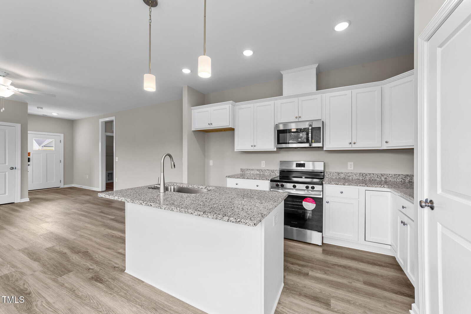 Modern kitchen with white cabinets, granite countertops, stainless steel appliances, a central island with a sink, and pendant lighting. The space features light wood flooring and neutral-colored walls. Entryway is visible in the background.